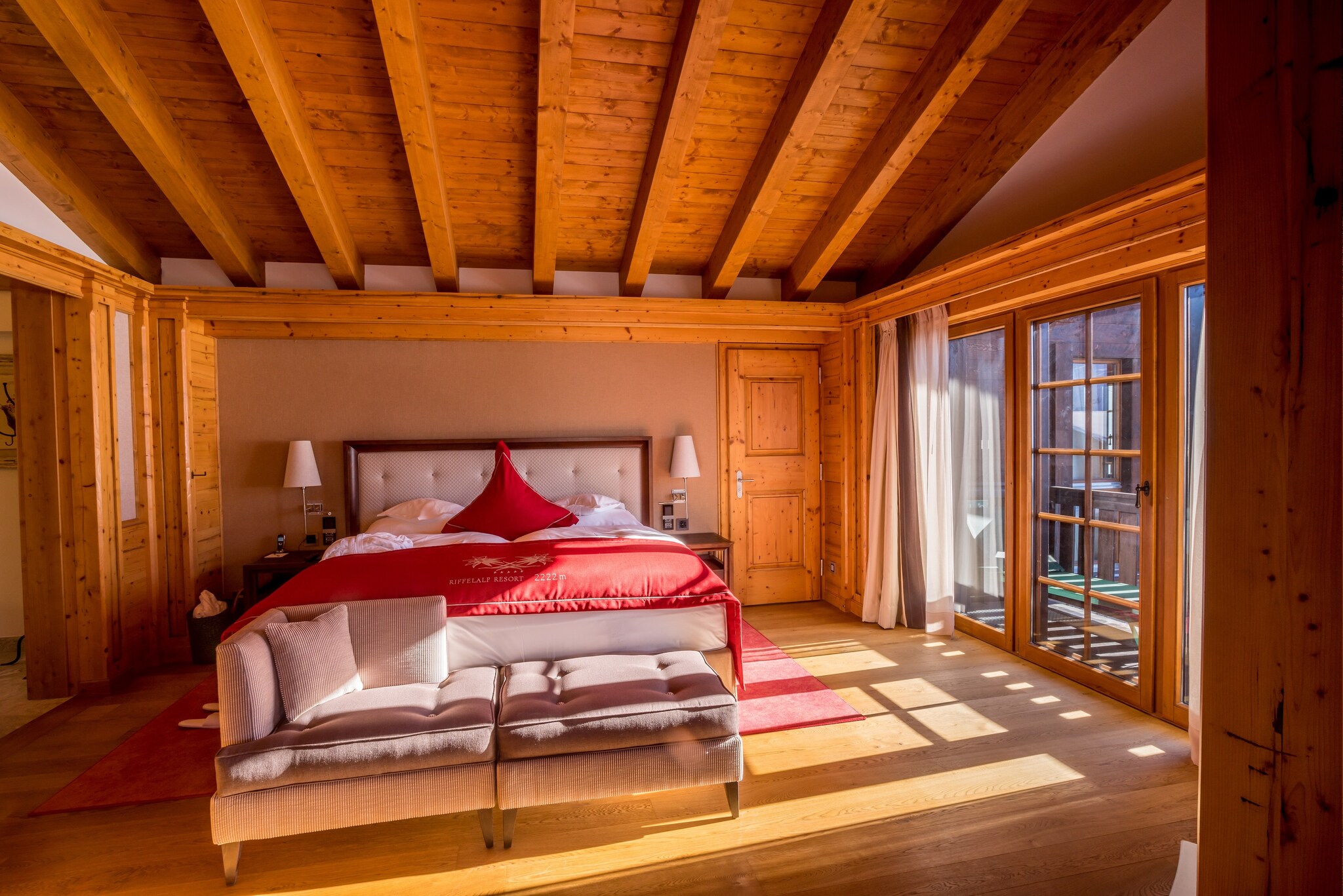 The sleeping room of a suite is paneled with knotted wood. A large white bed is accented with a red duvet cover and red triangular pillow.
