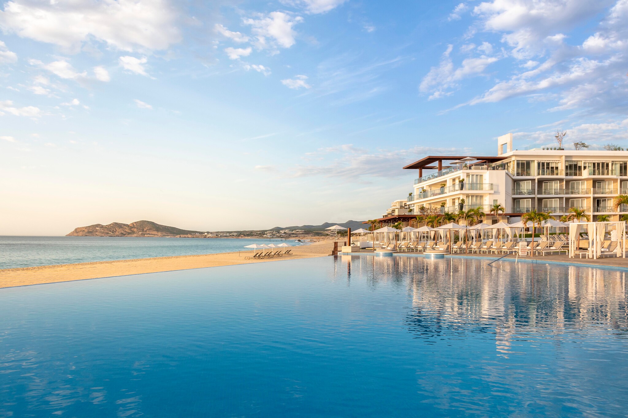 At Le Blanc Spa Resort Los Cabos, a large infinity pool abuts the beach on one side, with cabanas and white umbrellas lining the other side.