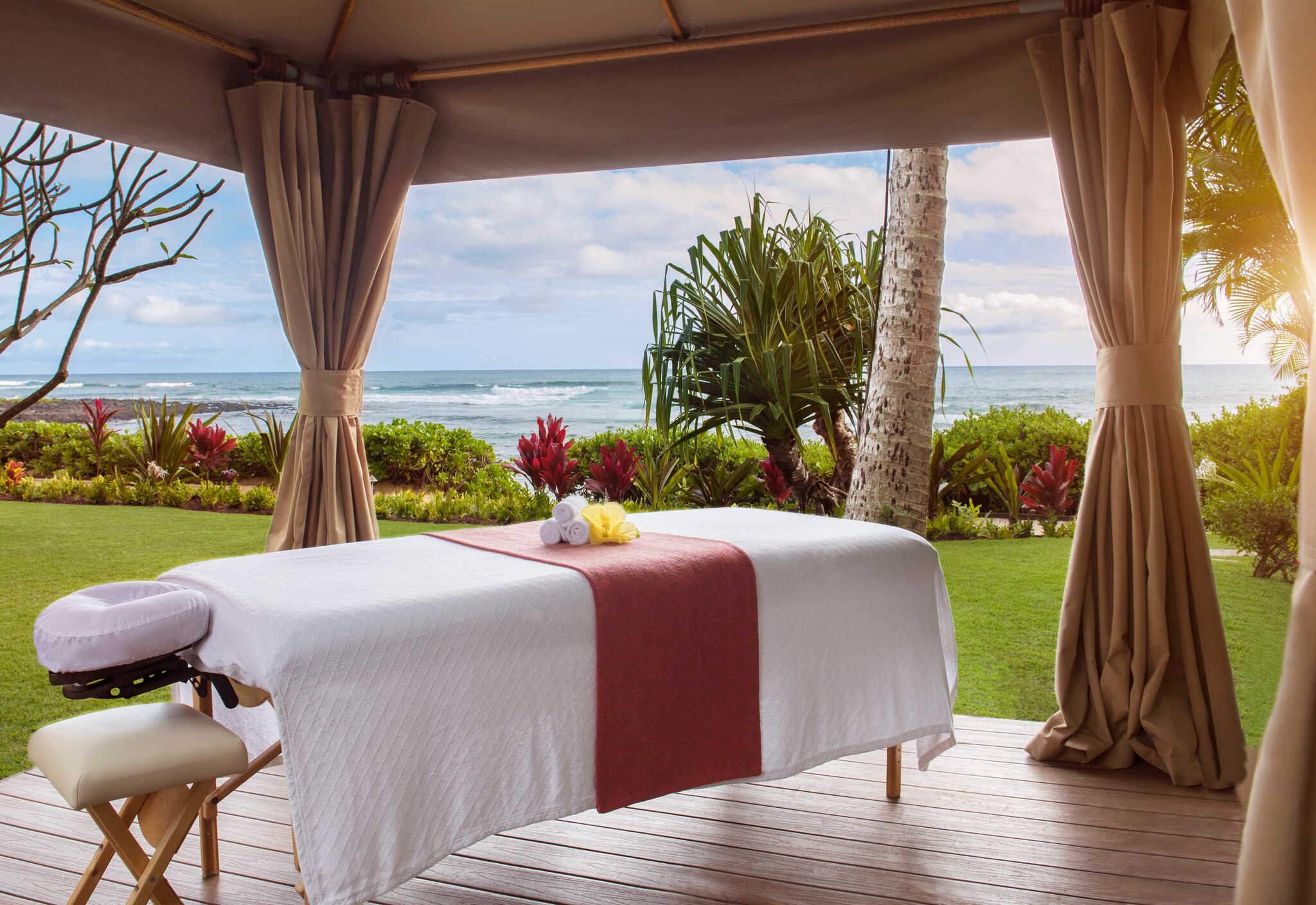 On a deck separated from the beach by beige curtains, a massage bed for The Spa at Ko’a Kea is arrayed with towels and a flower.