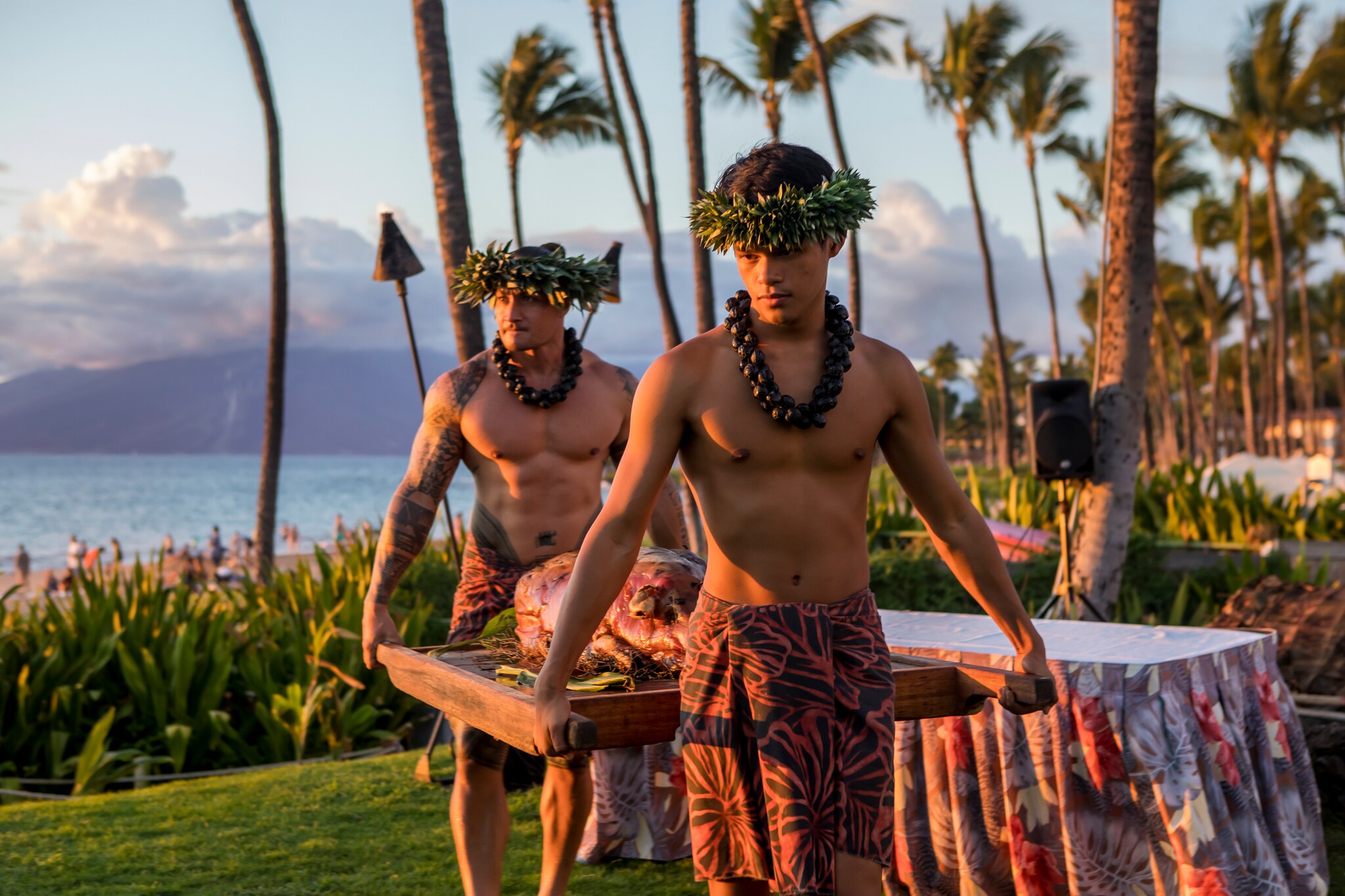 Two Hawaiian men in thick, kukui nut lei, and patterned sarongs with leafy lei po’o on their heads carry a roast pig into a lūʻau.