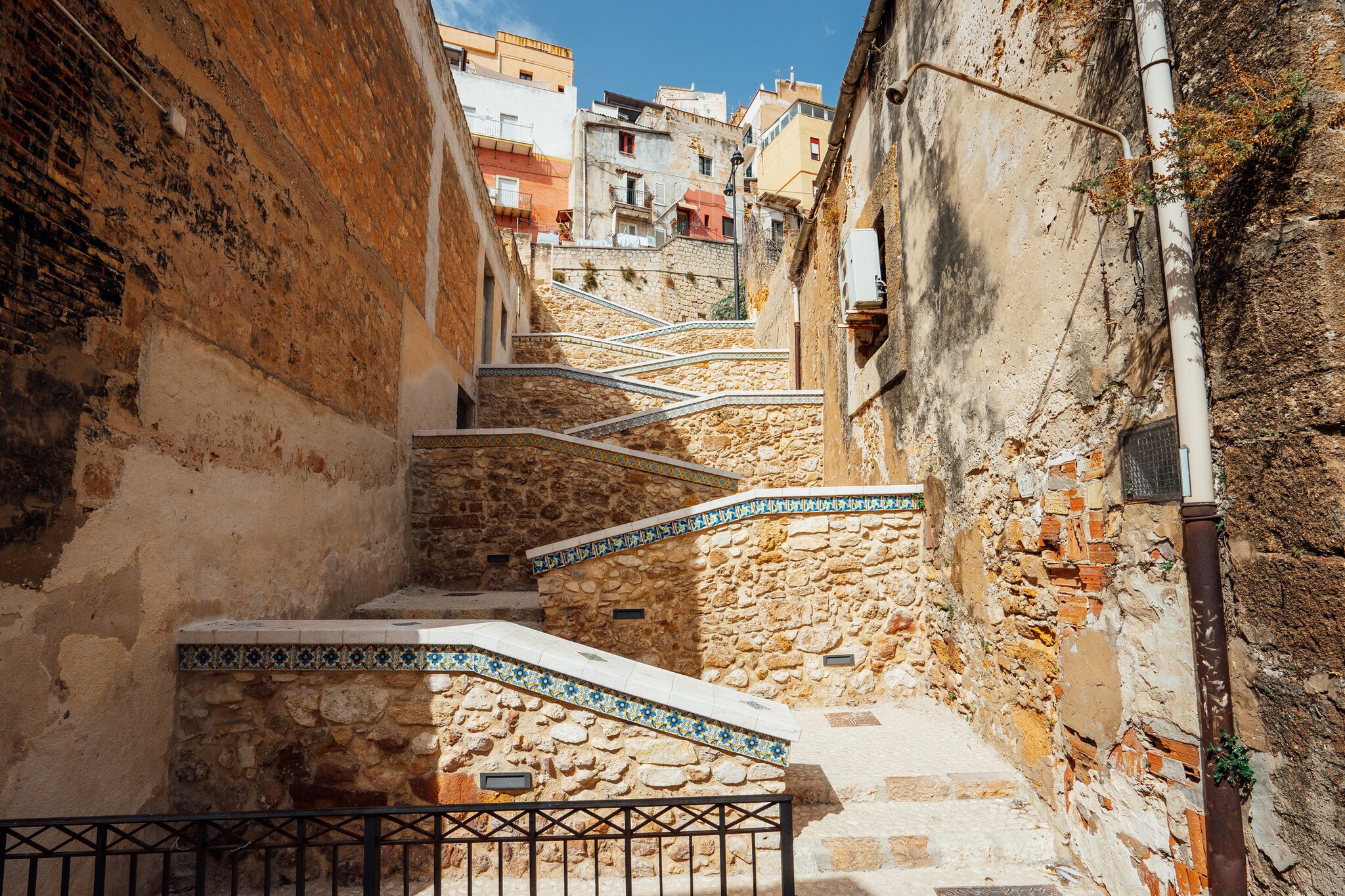 View up a stairway cutting steeply back and forth between two buildings. Blue and white majolica tiles line the tops of the handrails.