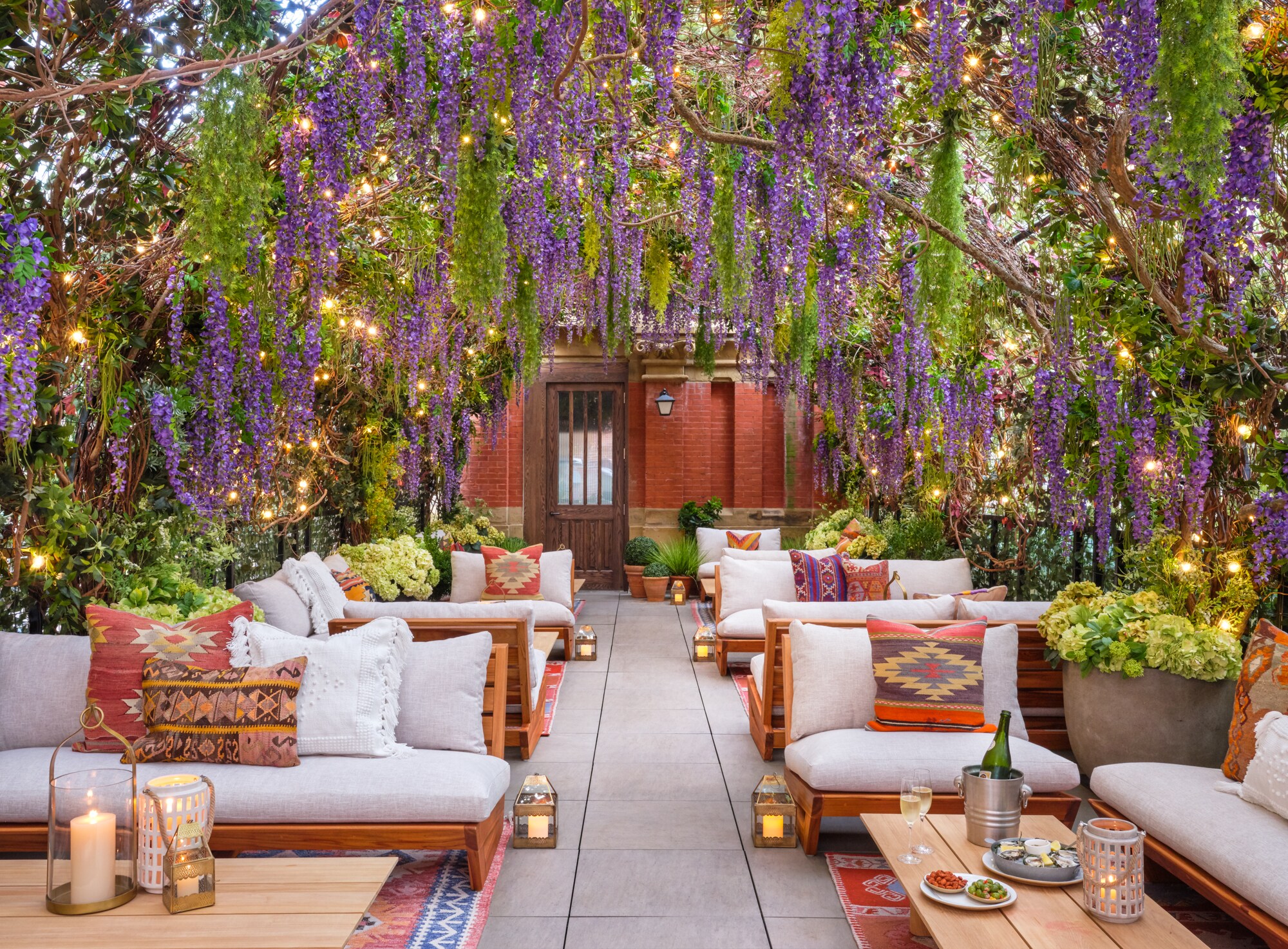 A penthouse terrace features rows of couches and chairs under archways vined with green leaves and purple flowers.