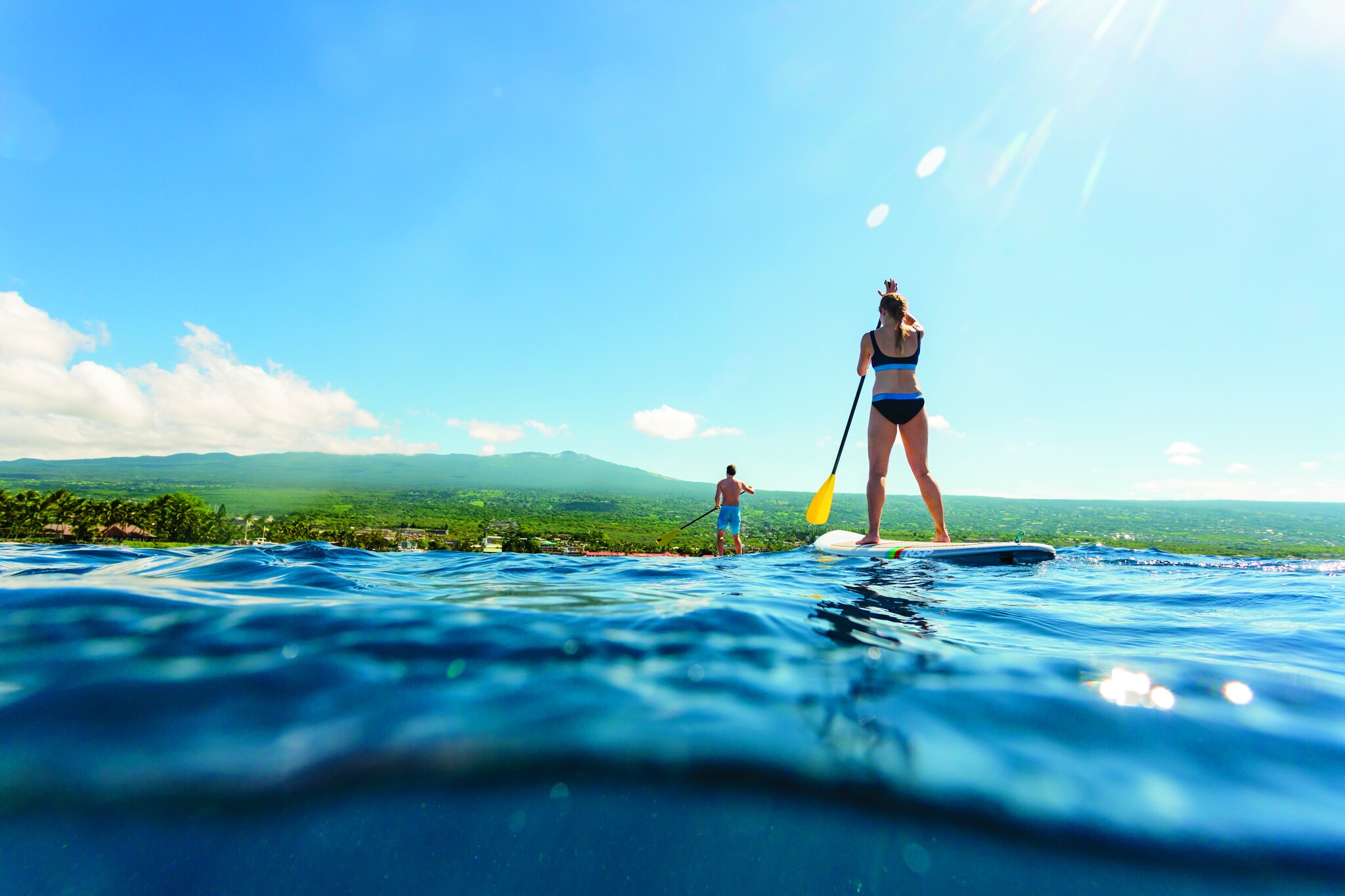 Water-level view of a white woman and a man in bathing suits paddle boarding towards a lush island.