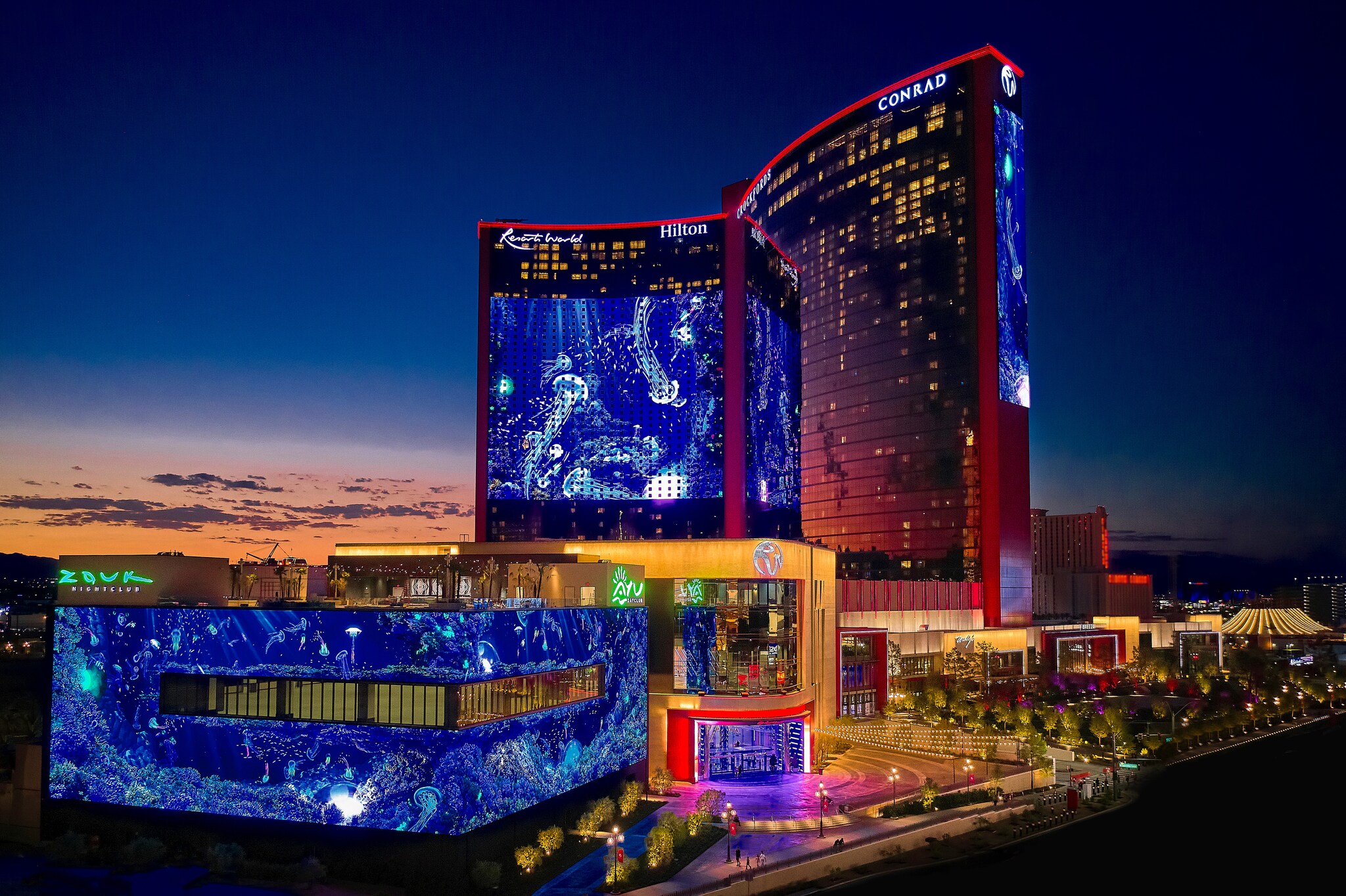 Aerial view of Resorts World Las Vegas at night. Bright blue undersea video is projected onto the exterior.