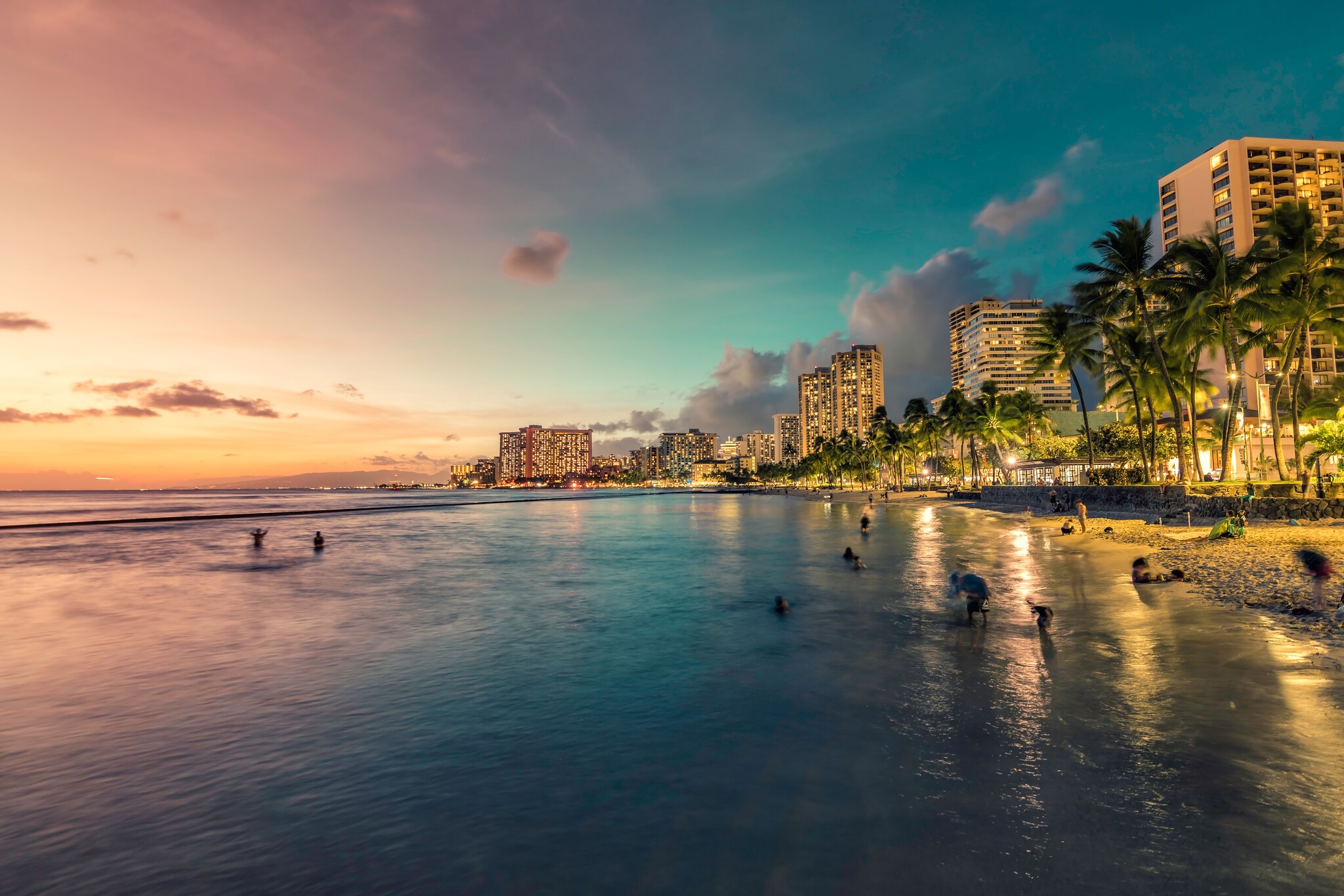 Waikiki beach at sunset. Beachgoers splash in the pink-tinged waters and the palms and skyscrapers down the coast are lit up for the night.