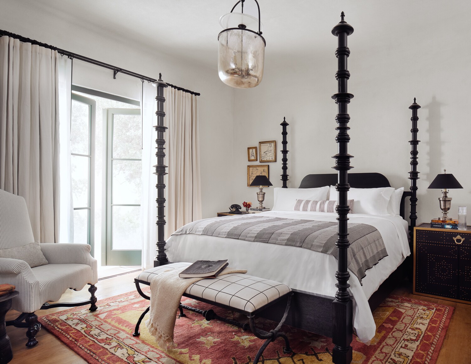 A room at the property features a four-poster bed, armchair, ottoman, and doors opening onto a sunny exterior.