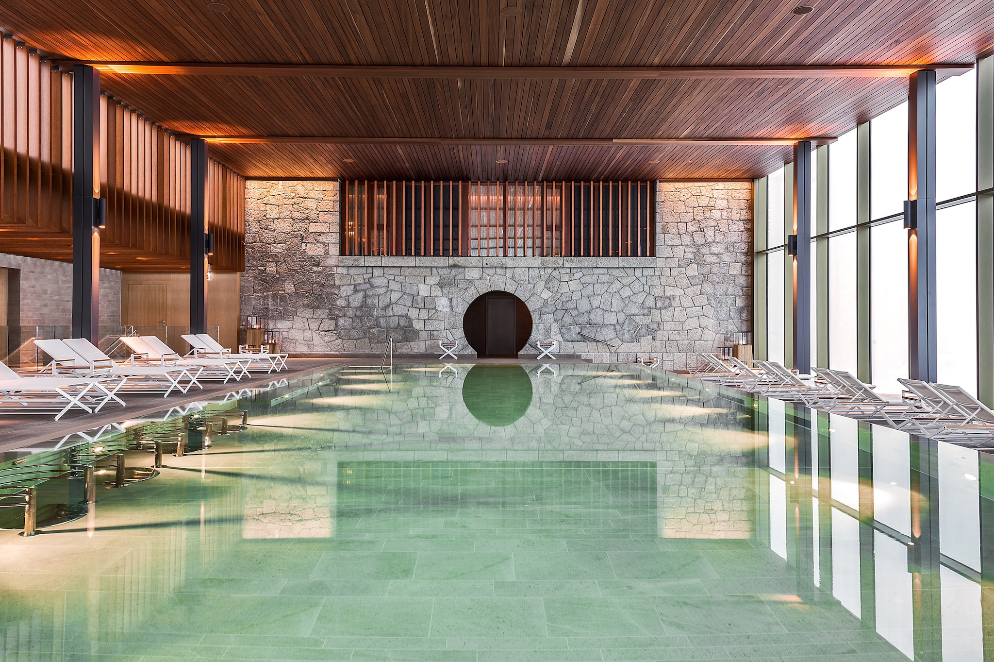An indoor pool is set in a high-ceilinged room with a wall of floor-to-ceiling windows, a wood-paneled ceiling, and rows of sun loungers.