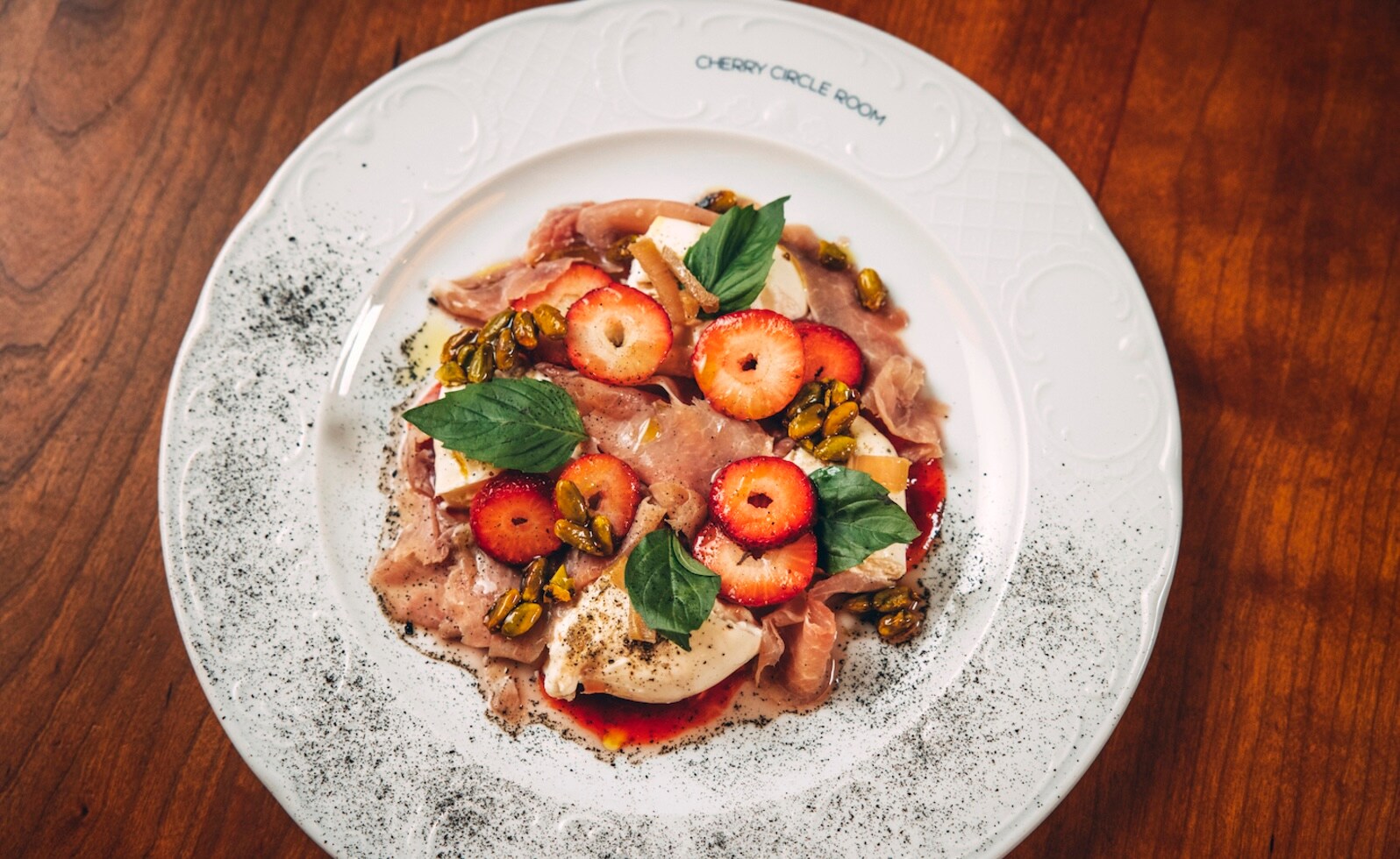 Overhead view of burrata with prosciutto, strawberry, rhubarb, and pistachio on a white dish stamped with the words Cherry Circle Room.