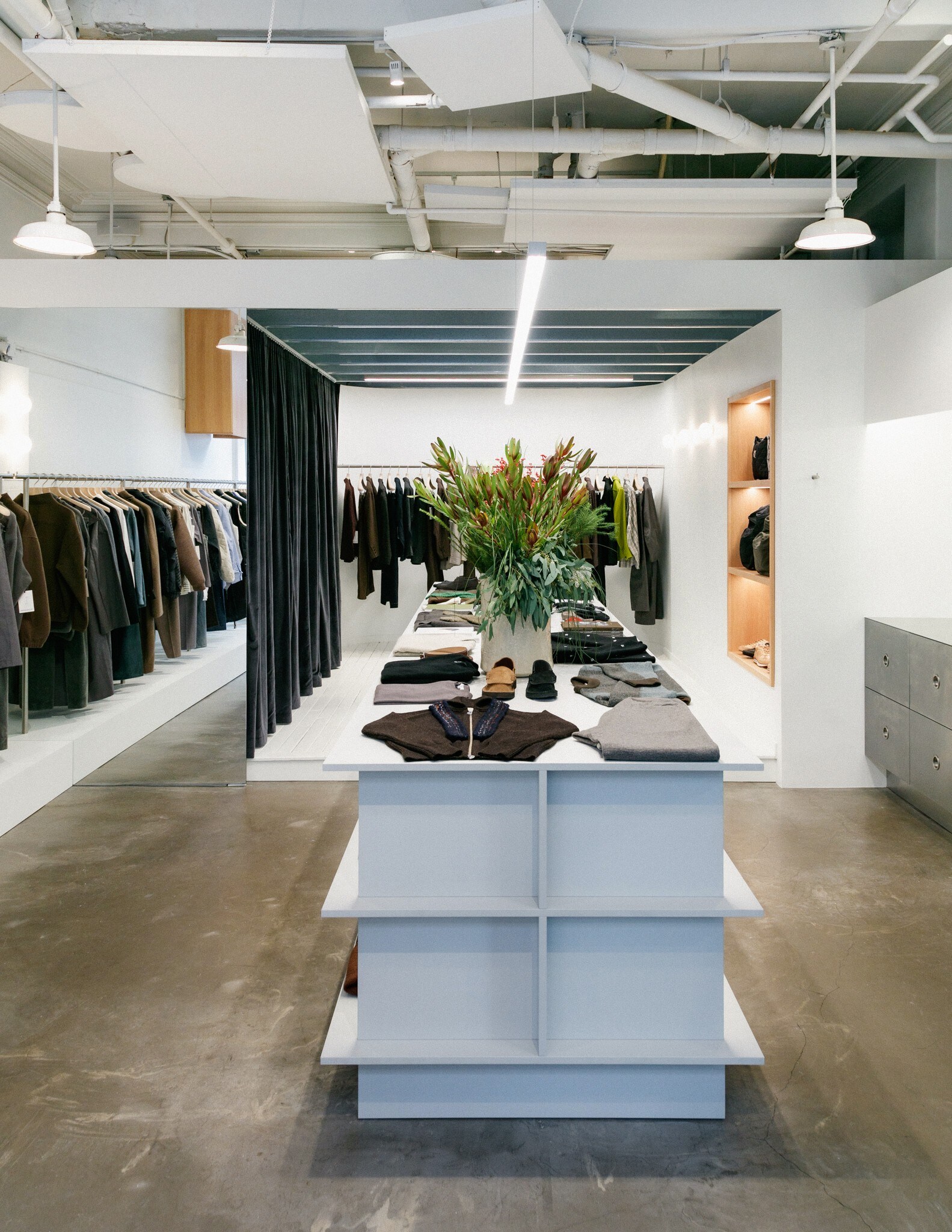 The white walls of a boutique are lined with long racks of clothing. A central table is set with clothes, shoes, and a large vase of flowers.