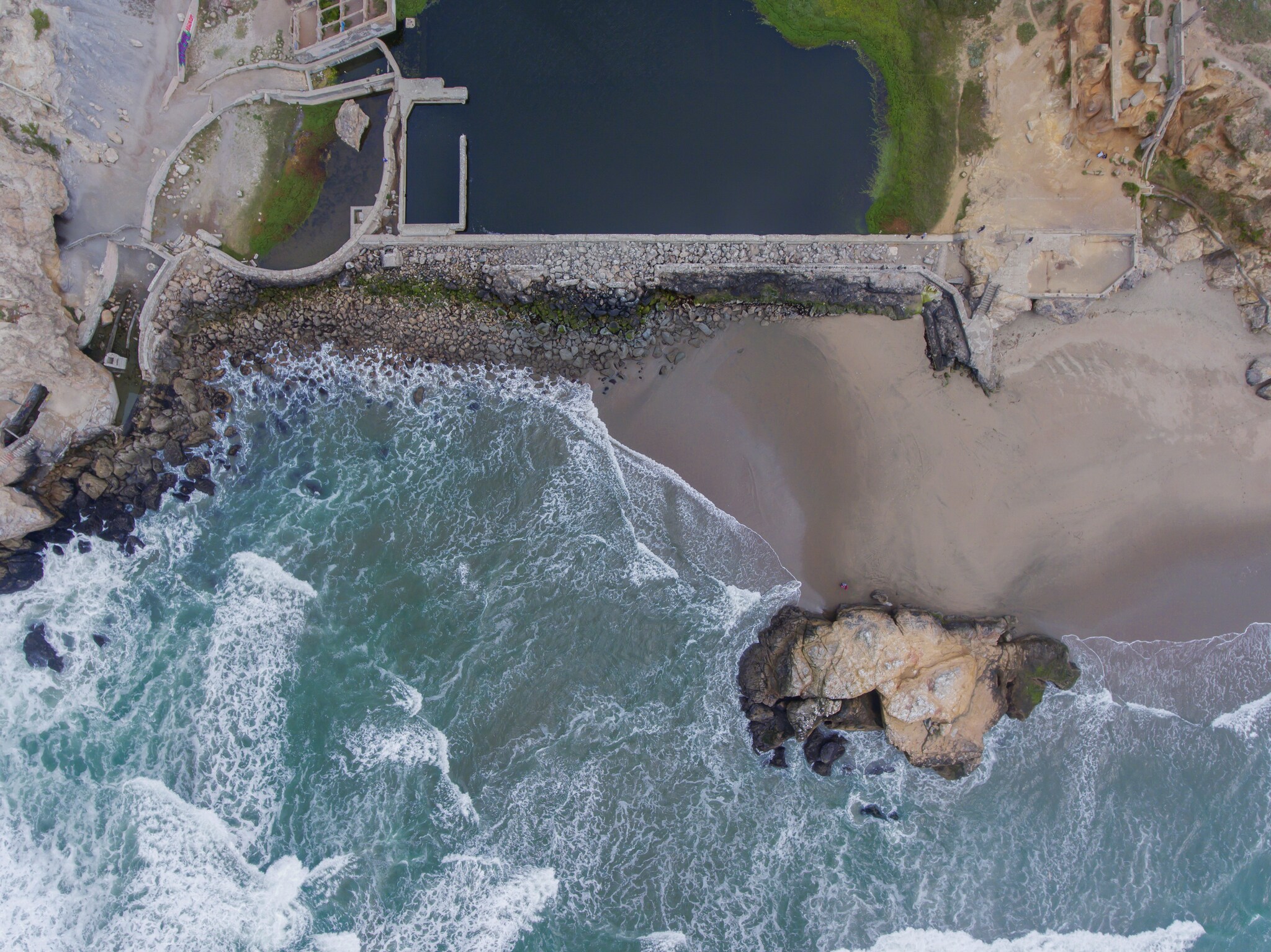 Overhead view of the beach adjoining a stone wall and the large, irregular blue expanse of the Sutro Baths.