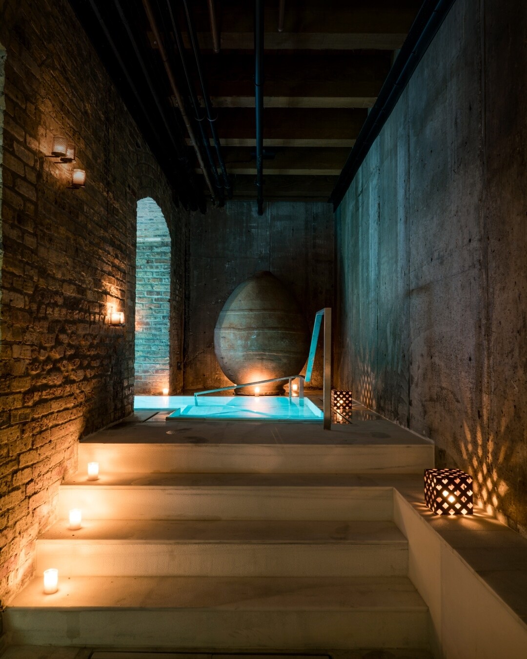  A dim stone-and-brick hallway leads to a softly glowing pool. Lights on the floor cast patterned shadows on the walls.