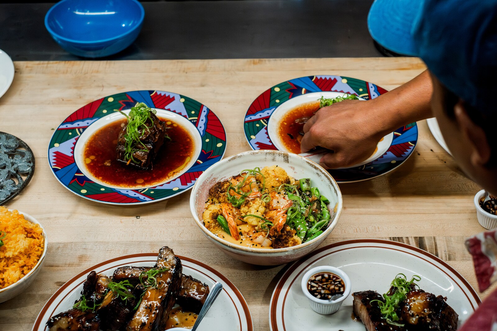 A brown-skinned chef reaches across a table of dishes to wipe sauce from the rim of a plate of meat surrounded by a dark red-brown broth.