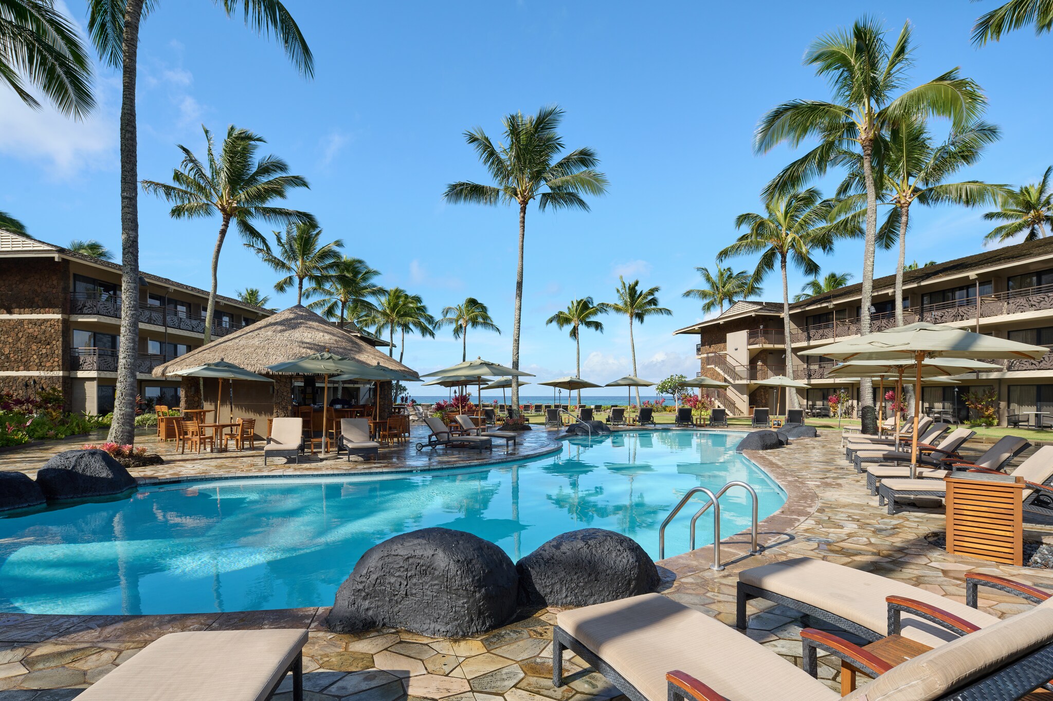The hotel pool is roughly bean-shaped, surrounded by low sun beds, umbrellas, palm trees, and a thatched-roof pool bar.