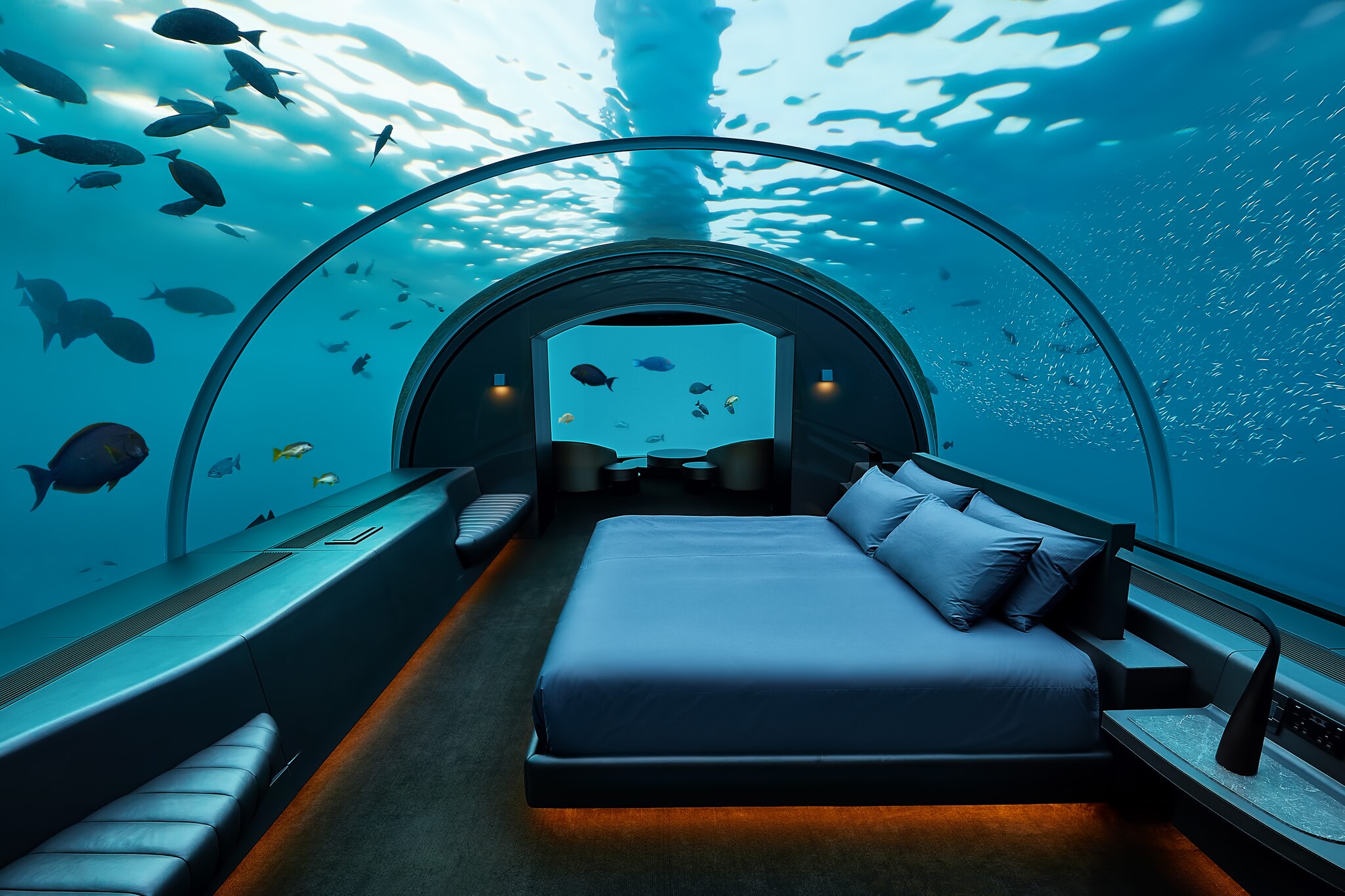 The underwater bedroom in the Muraka Residence has a wide bed with sleek blue sheets under an acrylic dome revealing fish swimming around.