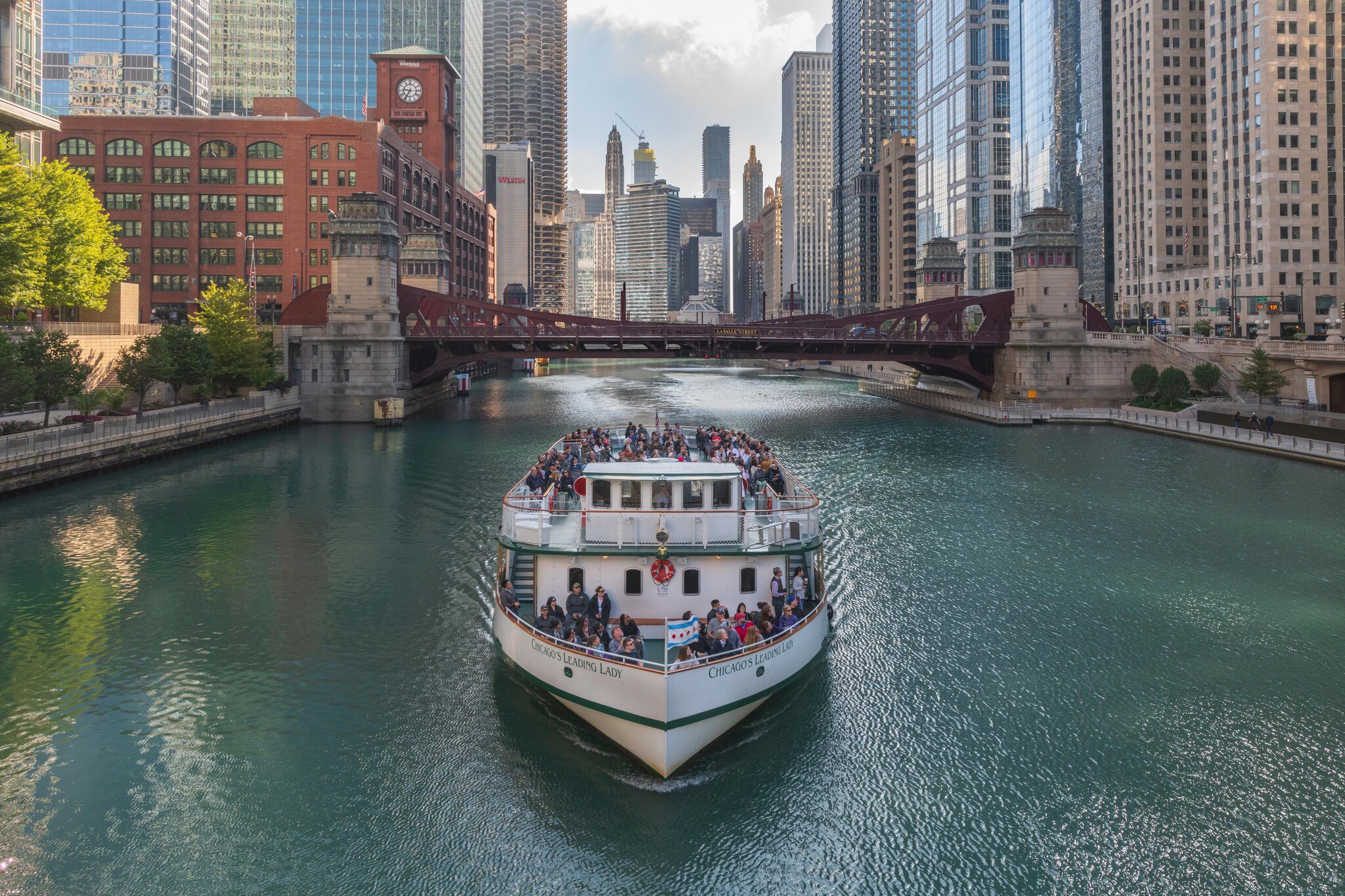 A white tour boat sails toward the viewer on the Chicago River, having just passed the Reid Murdoch Building and La Salle Street Bridge.