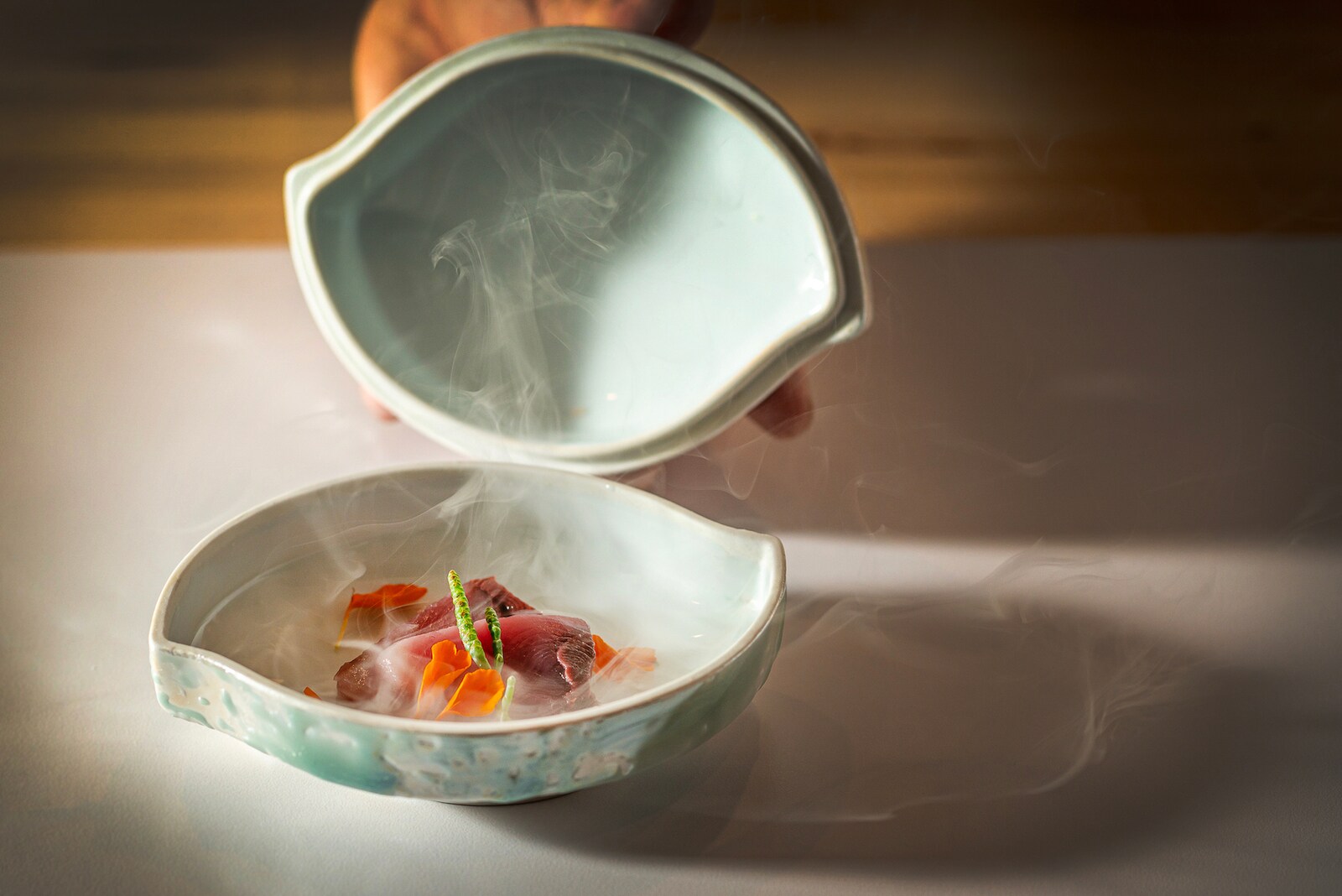A blue lemon-shaped ceramic cloche is lifted from a dish of red and orange curls, from which a white vapor curls up.