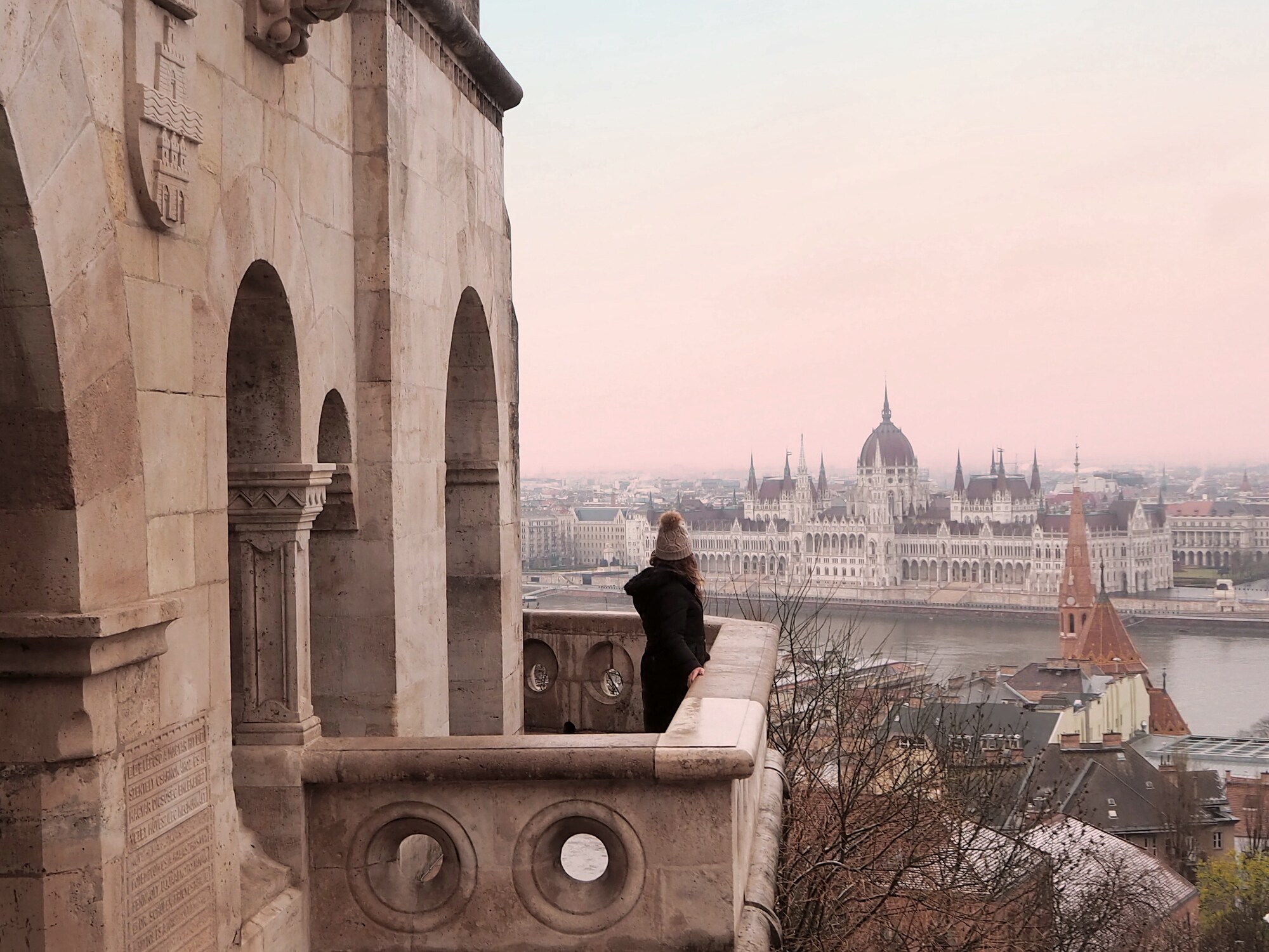 A woman in a knit cap with a pom-pom stands on a stone balcony and looks across the Danube at Budapest's parliamentary building.
