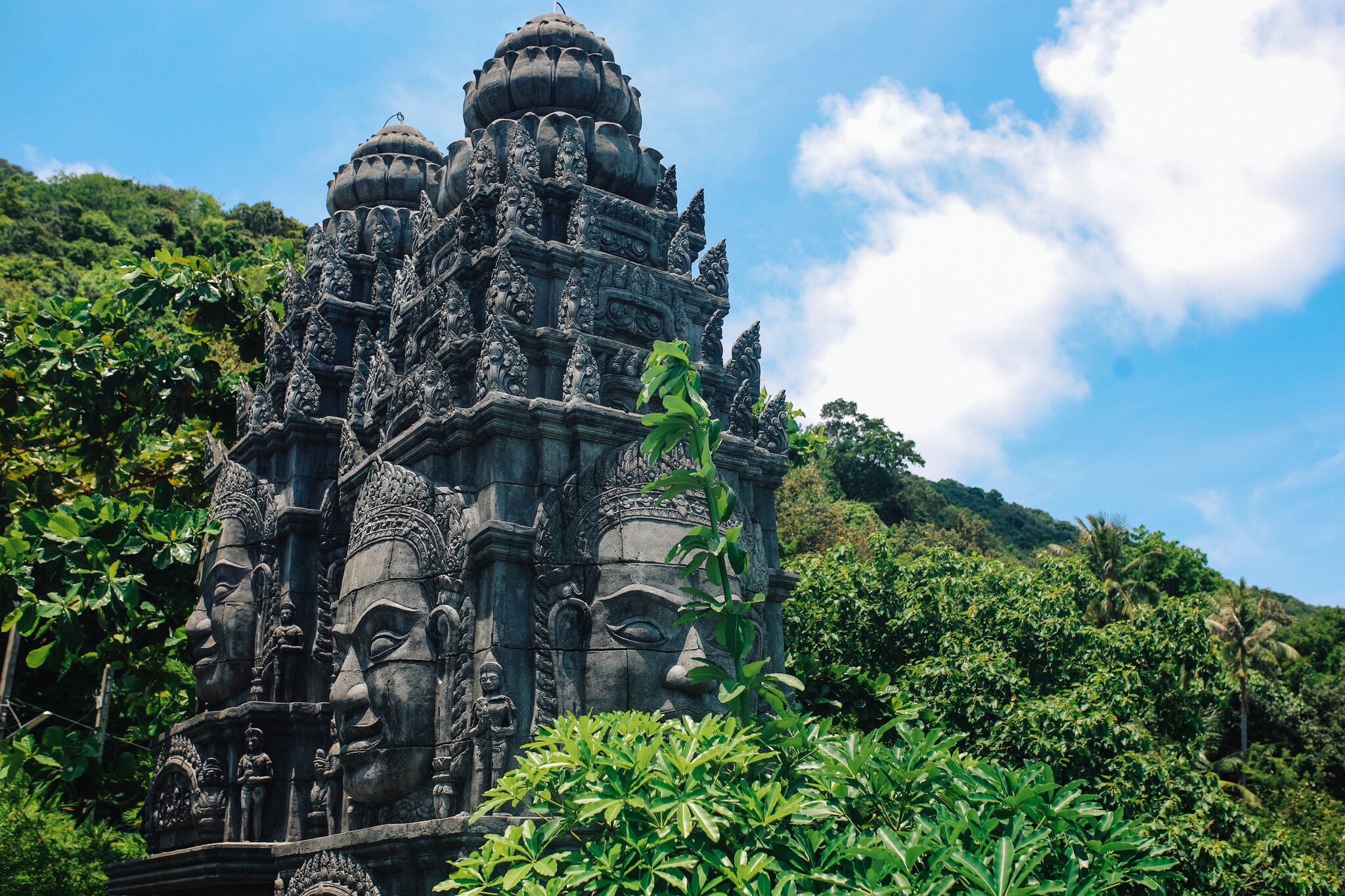 An elaborately carved dark gray stone temple, covered in Buddha faces and lotus motifs, rises out of lush greenery.