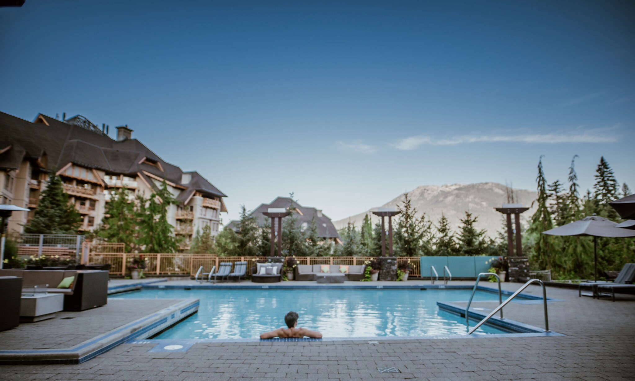 A man reclines in the pool with his elbows resting on the edge, looking across the water towards the trees and mountains beyond.