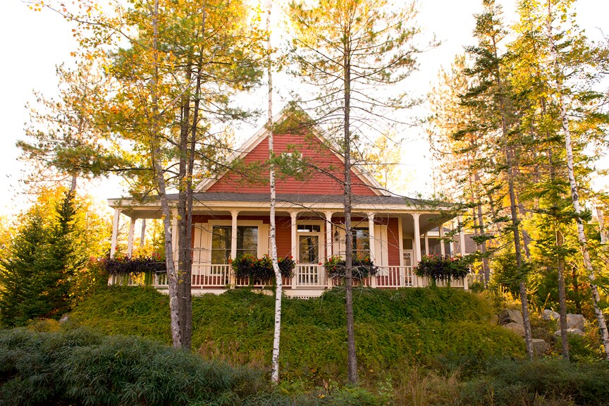 A red two-bedroom cottage with white trim sits atop a small hill, surrounded by trees. The top is steeply gabled, with a wide porch covering.
