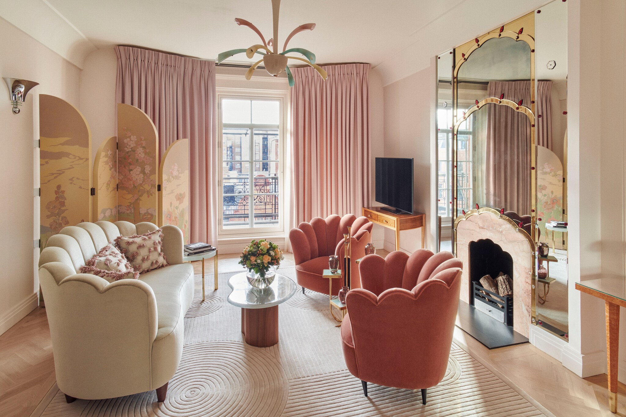 The Mayfair Suite is decorated pink and white, with mirrors around a fireplace, and a folding screen almost obstructing doors to a balcony.
