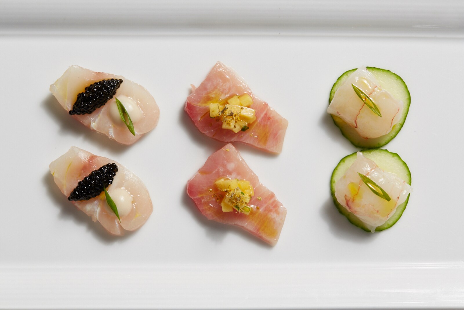 On a white dish, two rows of appetizers showcase fresh fish, cucumbers, and caviar in bright colors.