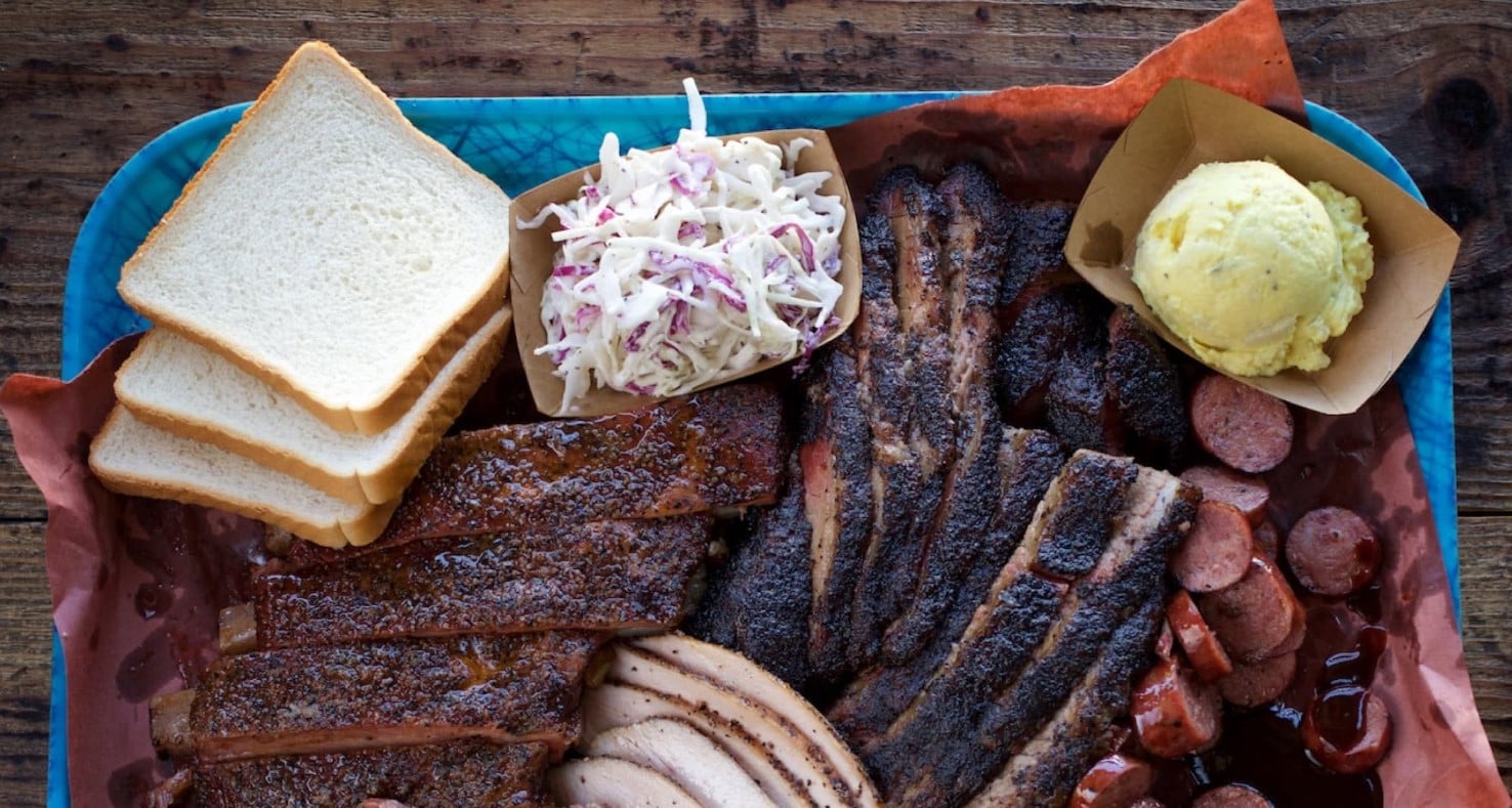 Overhead view of a tray laden with piles of finely seasoned and charred meat, slaw, slices of white bread, and a big scoop of mashed potatoes.