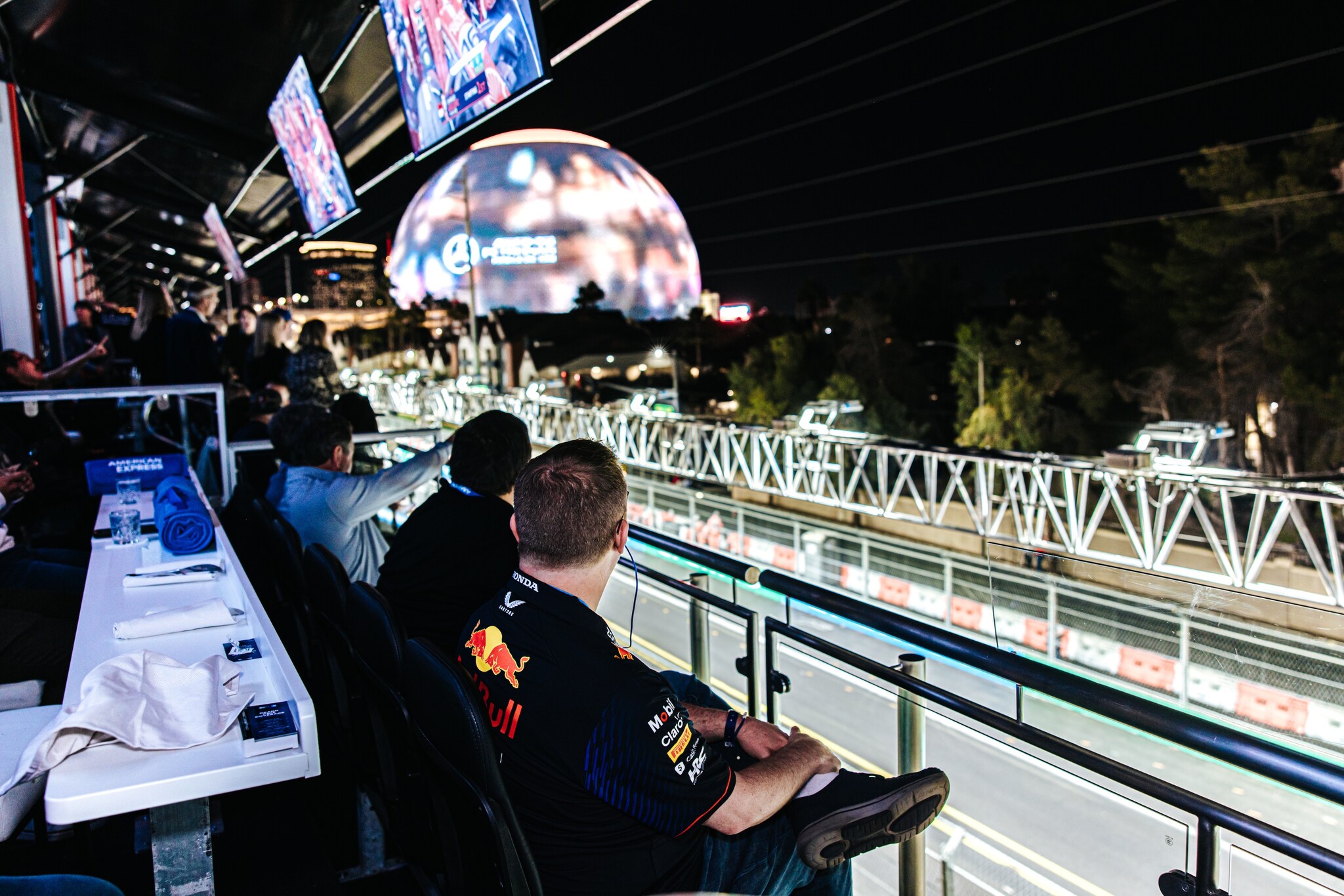 View from behind of a row of fans looking off the edge of the Amex Trackside Lounge at the brightly-lit track.
