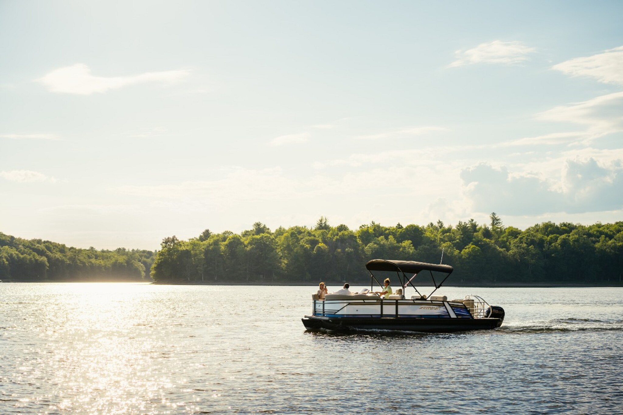 A small private boat motors across a lake in front of densely forested shores. Three people recline in the sun on the boat.