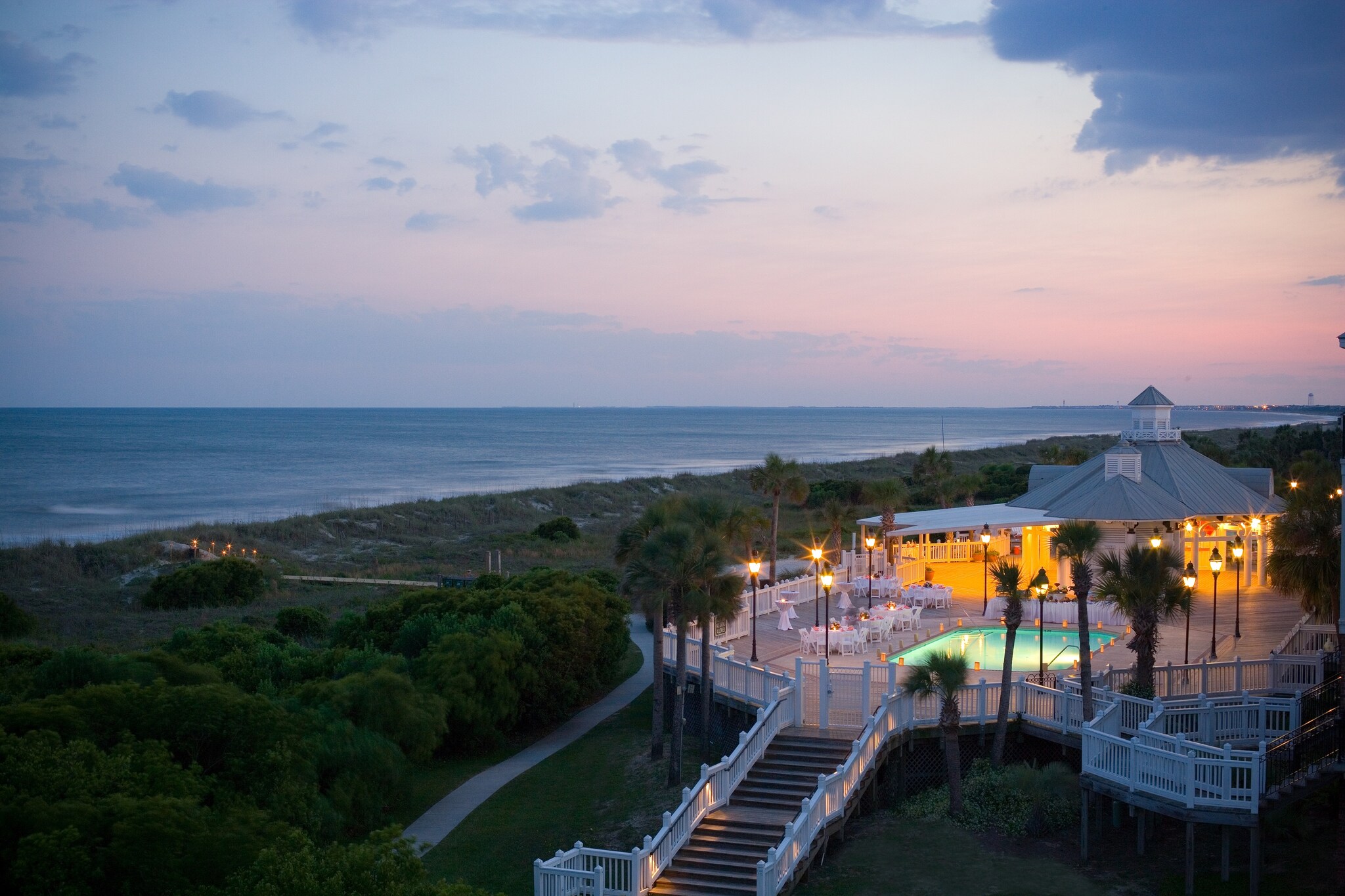 At sunset on the beach, a gazebo is lit up golden next to a pool and a deck set with white furniture. 