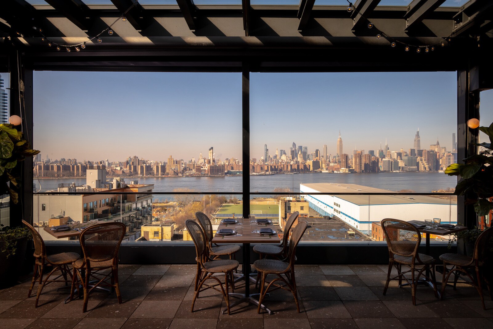 The indoor dining room is filled with light rattan chairs around small square tables. Floor-to-ceiling windows look towards the water.