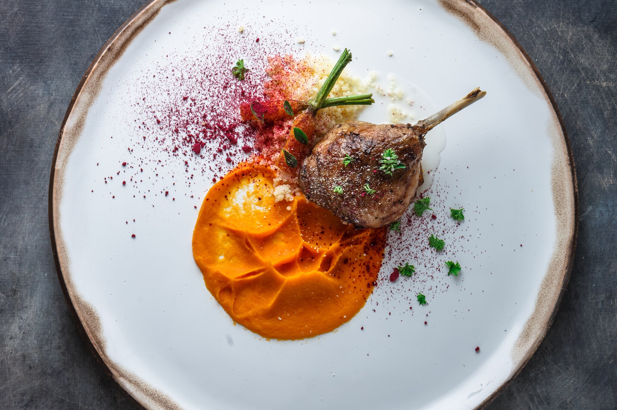 A bone-in cut of meat rests against small carrots and a bright orange paste. Colorful red and yellow dust sprinkles the dish.