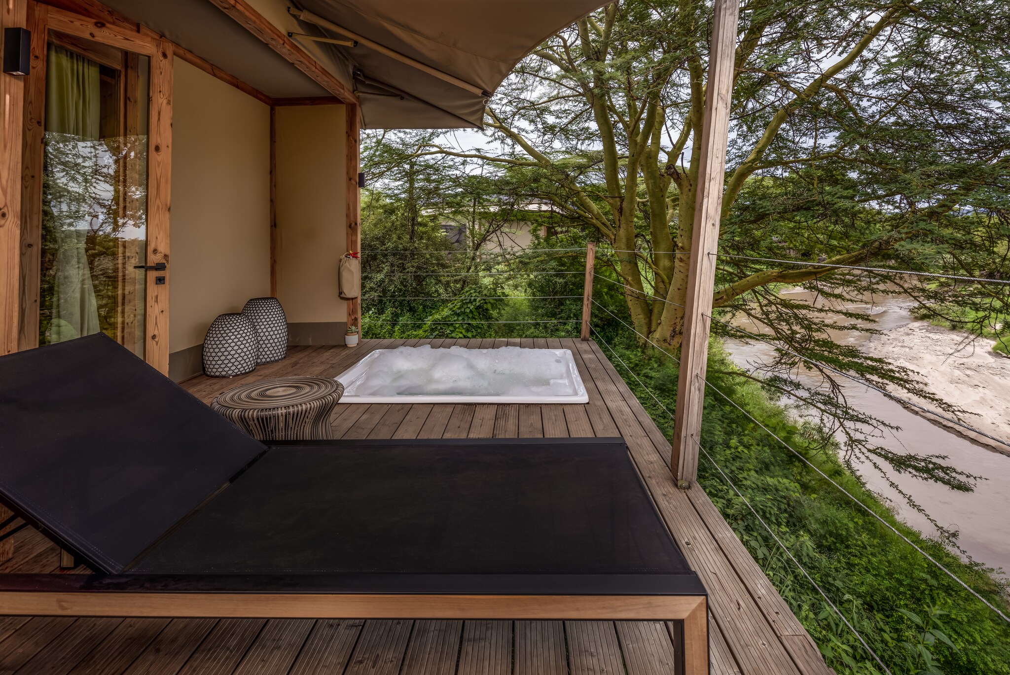A low black sun bed sits on a deck next to a bubbling whirlpool bath. Beyond, dense foliage and sand are visible.