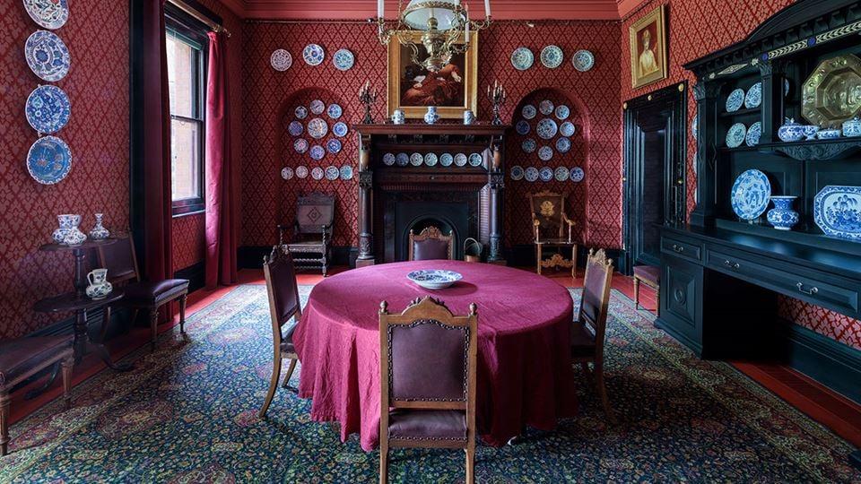  The dining room of the Leighton House Museum features Victorian décor, with patterned wallpaper, mounted china dishes, and a red table.
