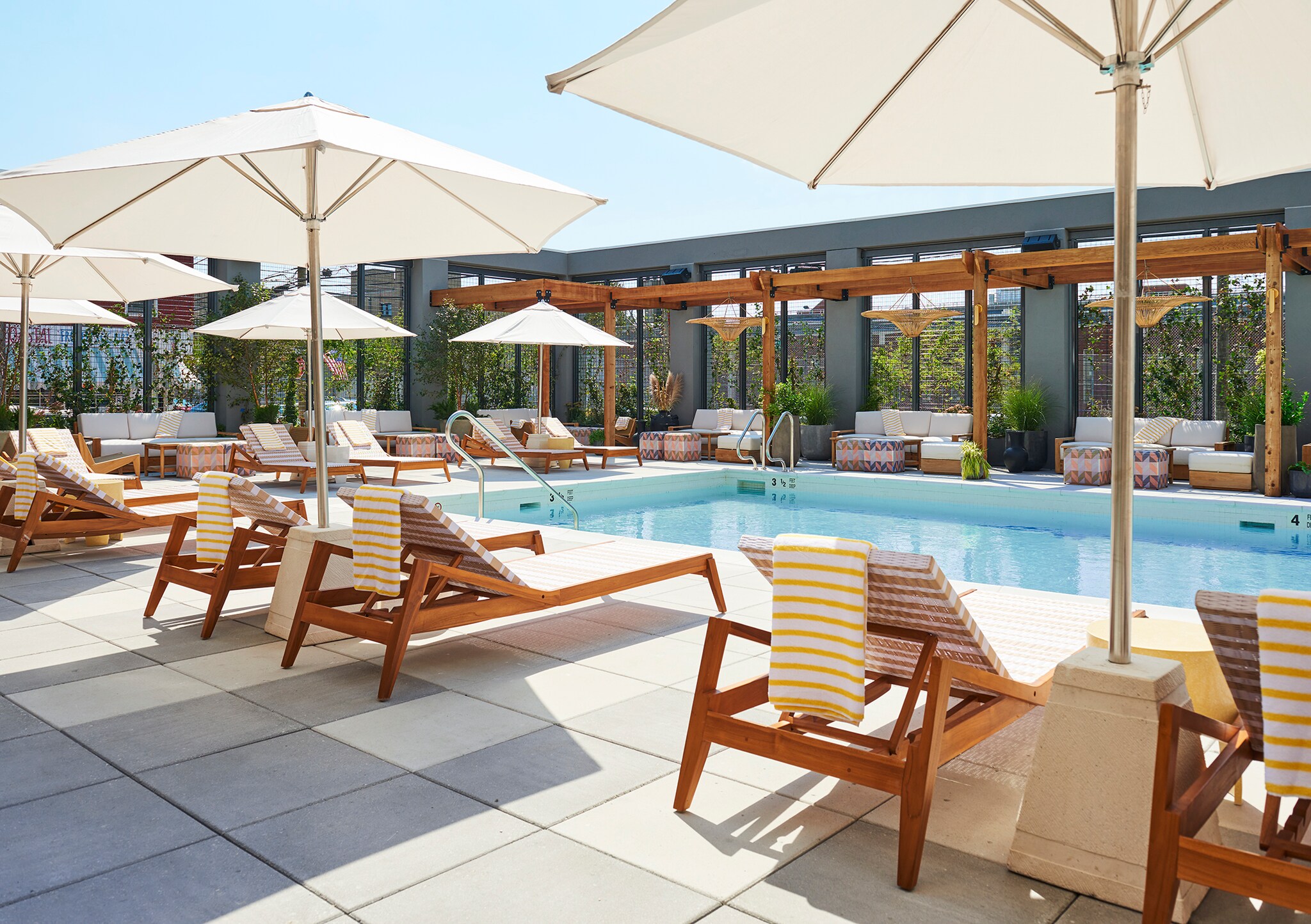 The hotel pool deck is paved with light gray tiles. Sun beds with striped towels are arrayed around the pool under white umbrellas.