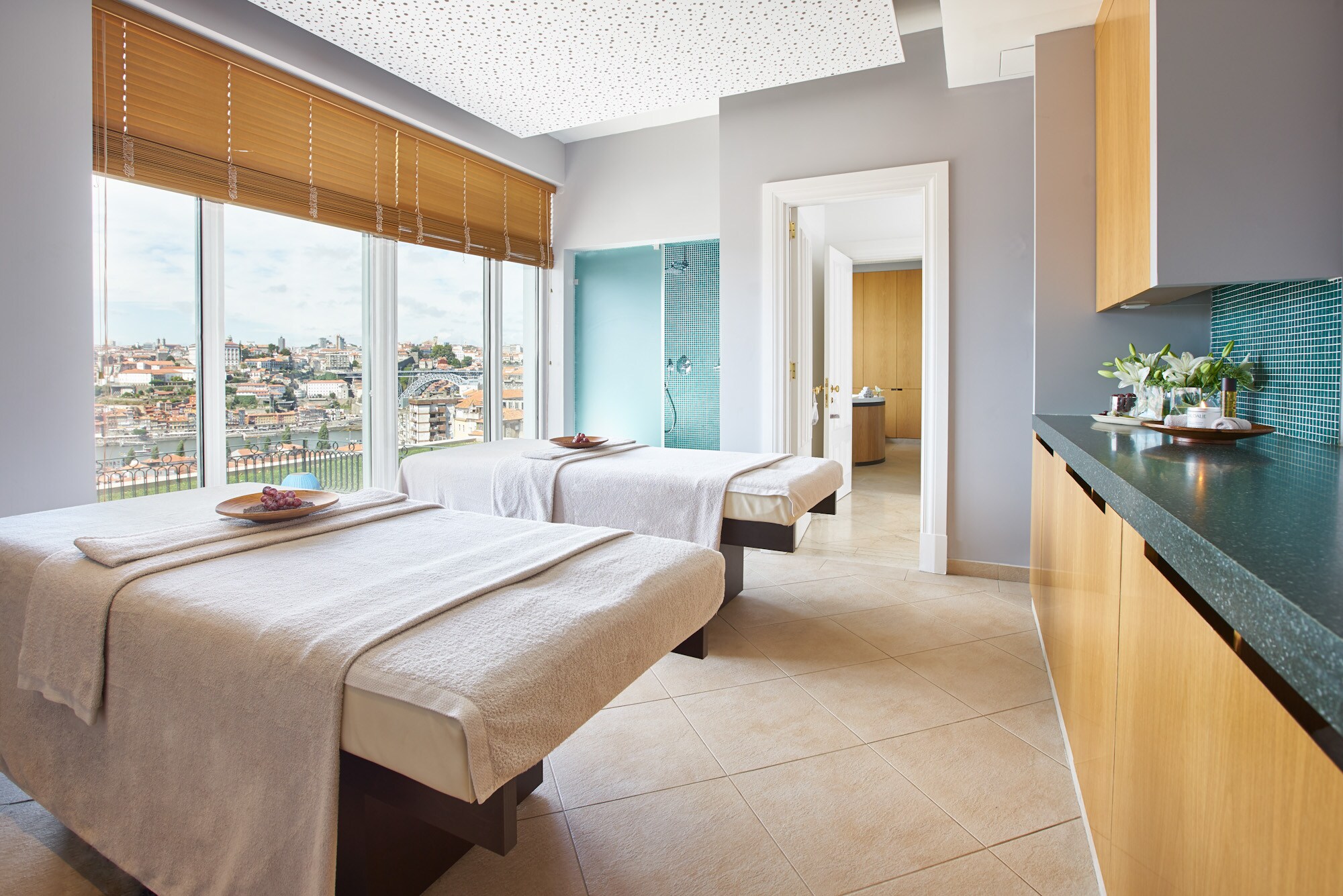 A bright spa treatment room has two massage beds laid out with towels and a tray of fruit, a blue-tiled shower, and tall windows.