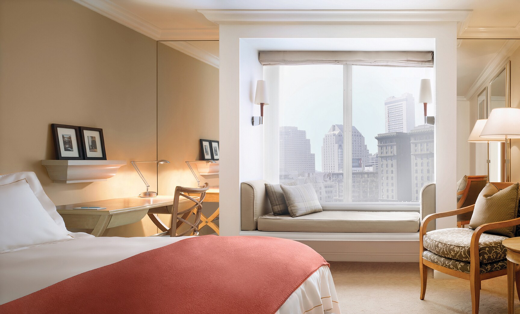 A Deluxe Stockton Street guest room at Taj Campton Place has tan walls, a red blanket at the foot of the bed, and a white boxed-off window seat.