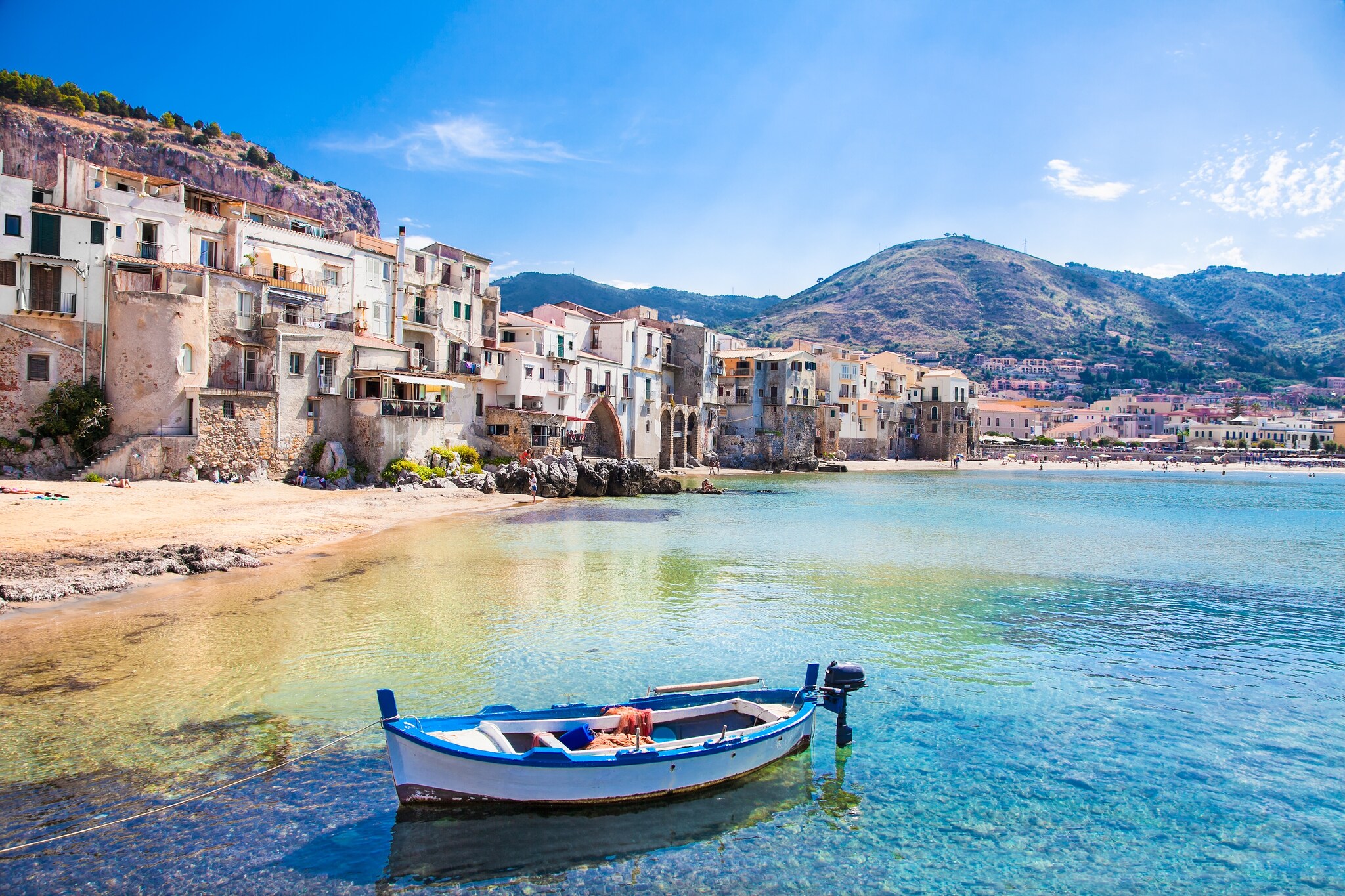 A white rowboat with blue trim is tethered in the shallows of a cove lined by rows of closely-packed stucco buildings.