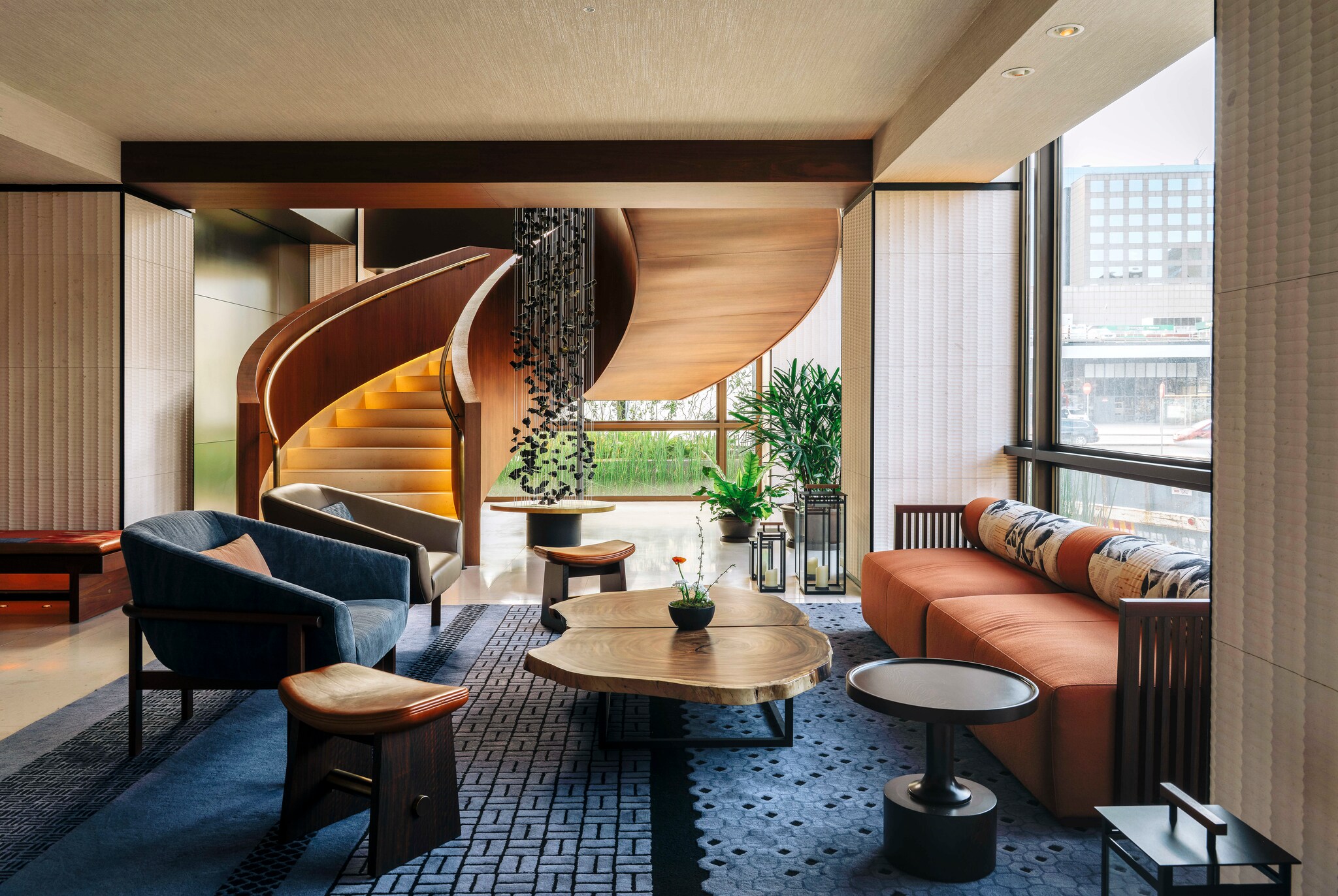 In Nobu Hotel Barcelona, a curved red-wood staircase winds down into a sitting area with a blue rug and mid-century modern furniture.