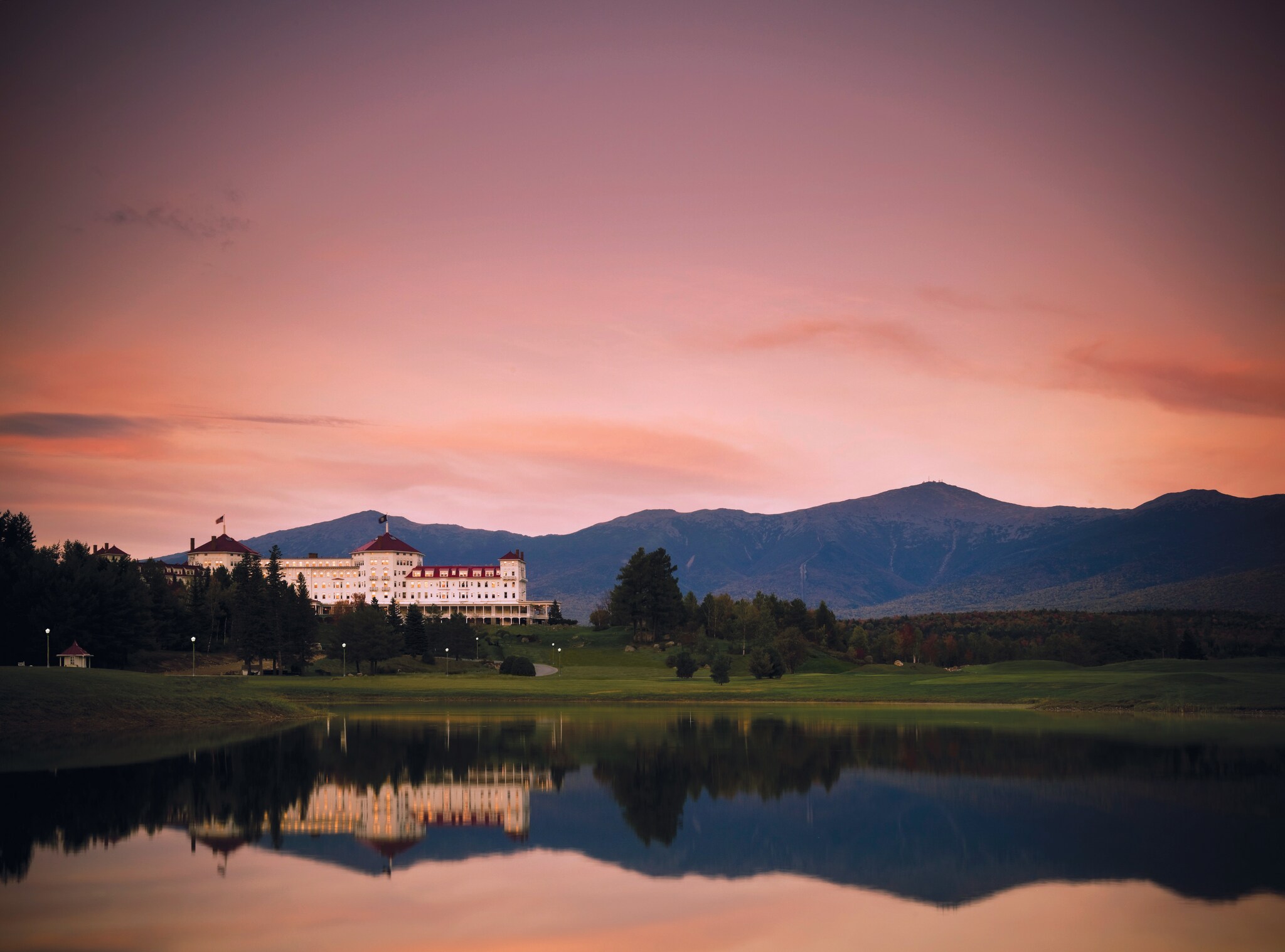 Across a glassy lake, on a forested hill, the hotel is a grand white building with a red roof under a sunset sky in front of blue mountains.