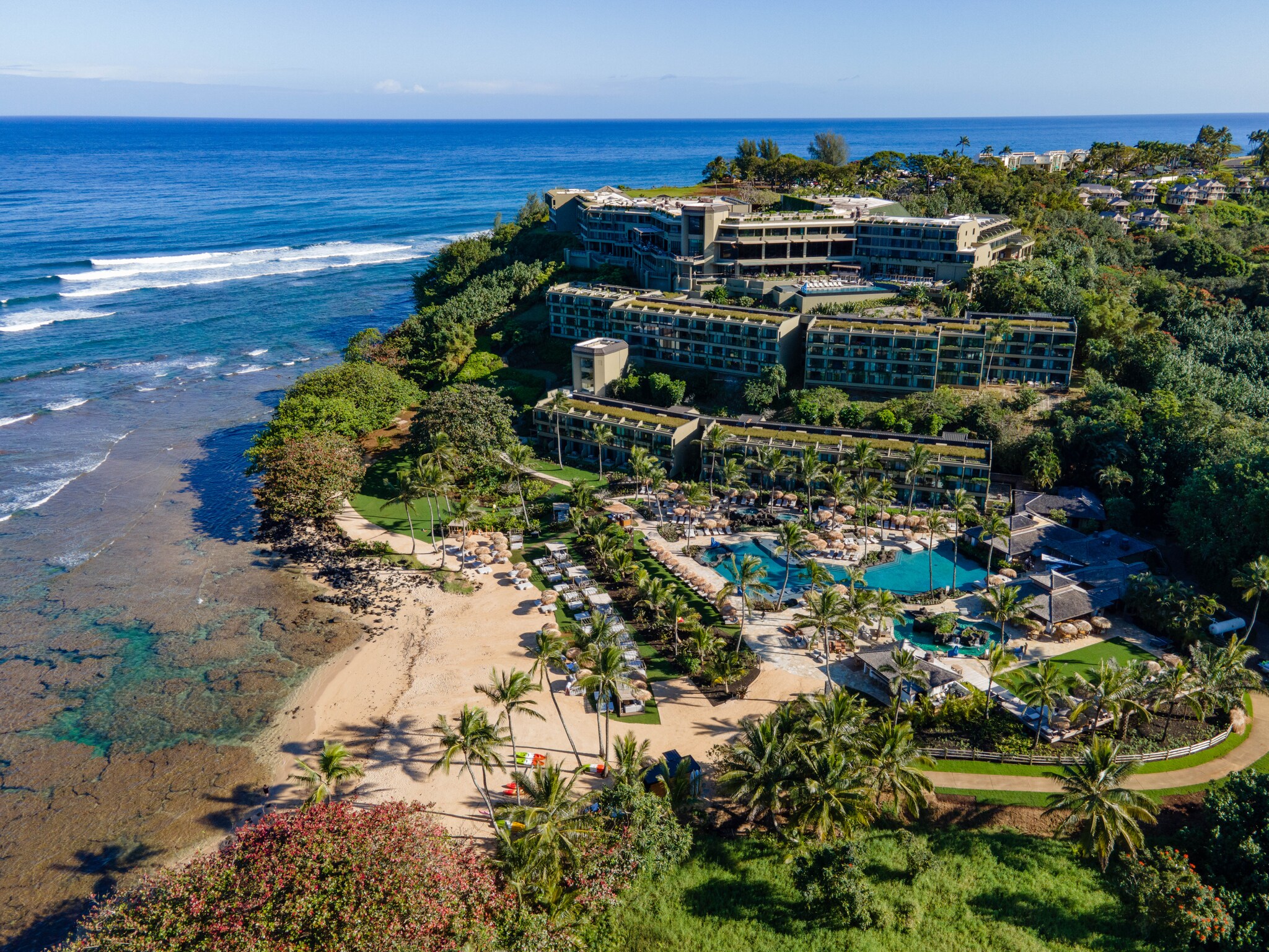 Aerial view of 1 Hotel Hanalei Bay. Multi-story buildings are built up the side of a hill, with a pool, cabanas, and a beach at the bottom.
