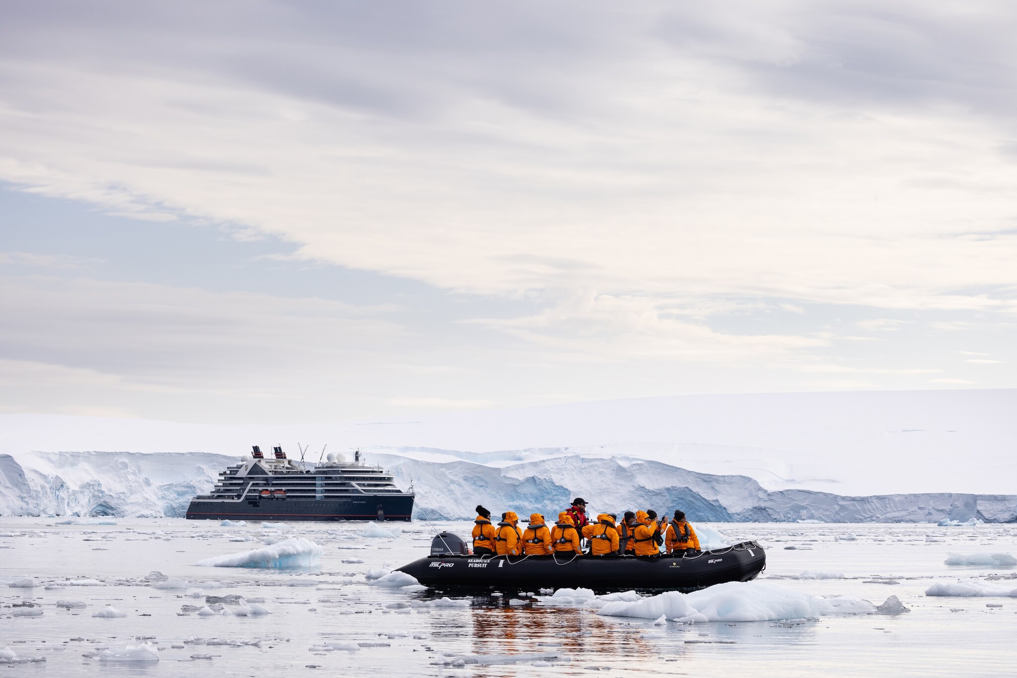 An inflatable boat full of people in orange jackets looks around at floating ice. Beyond, a Seabourn ship floats in front of a snowy shore.