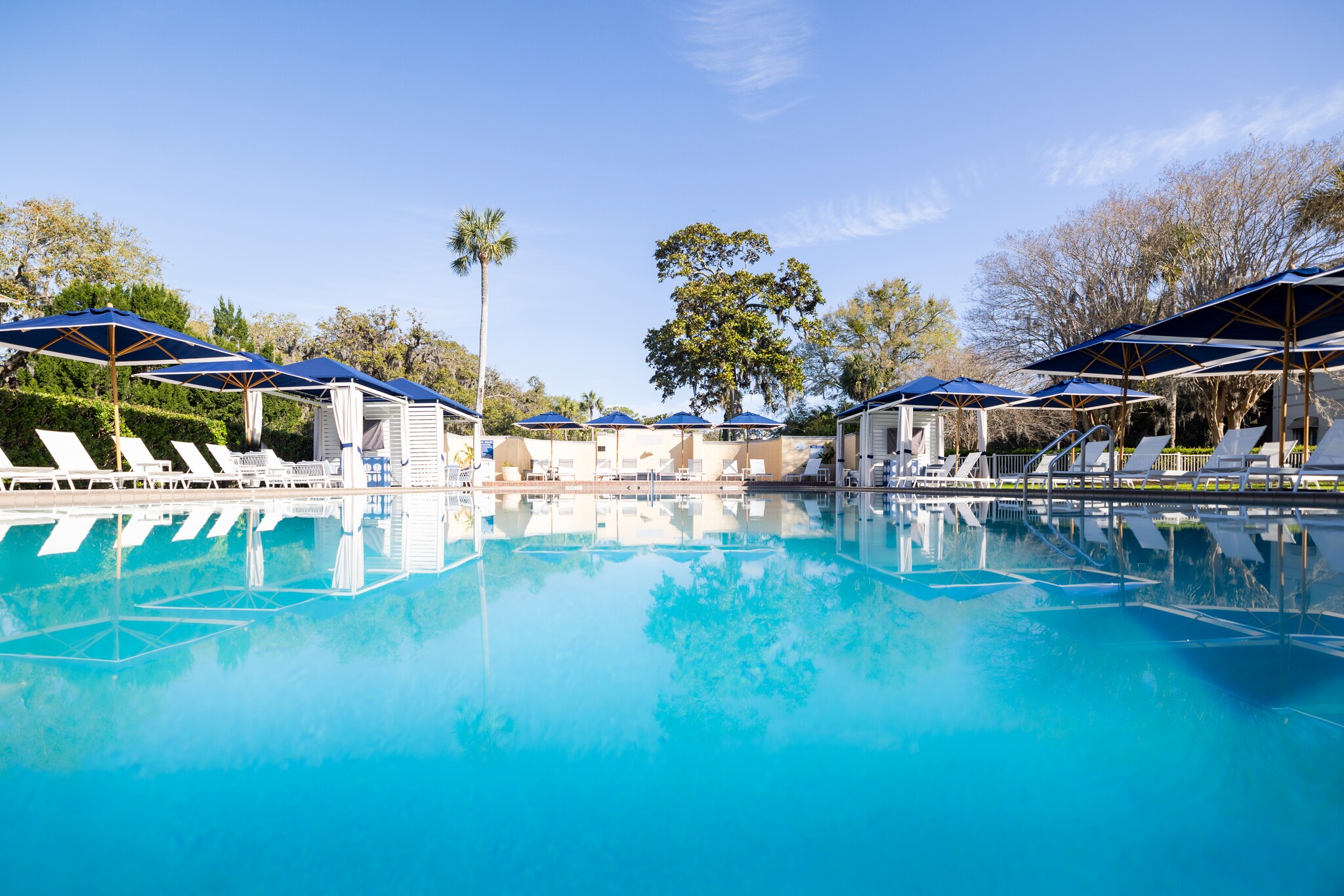 The resort pool is bright, and white sun beds and square blue umbrellas line the pool on either side, with hedges and trees behind.