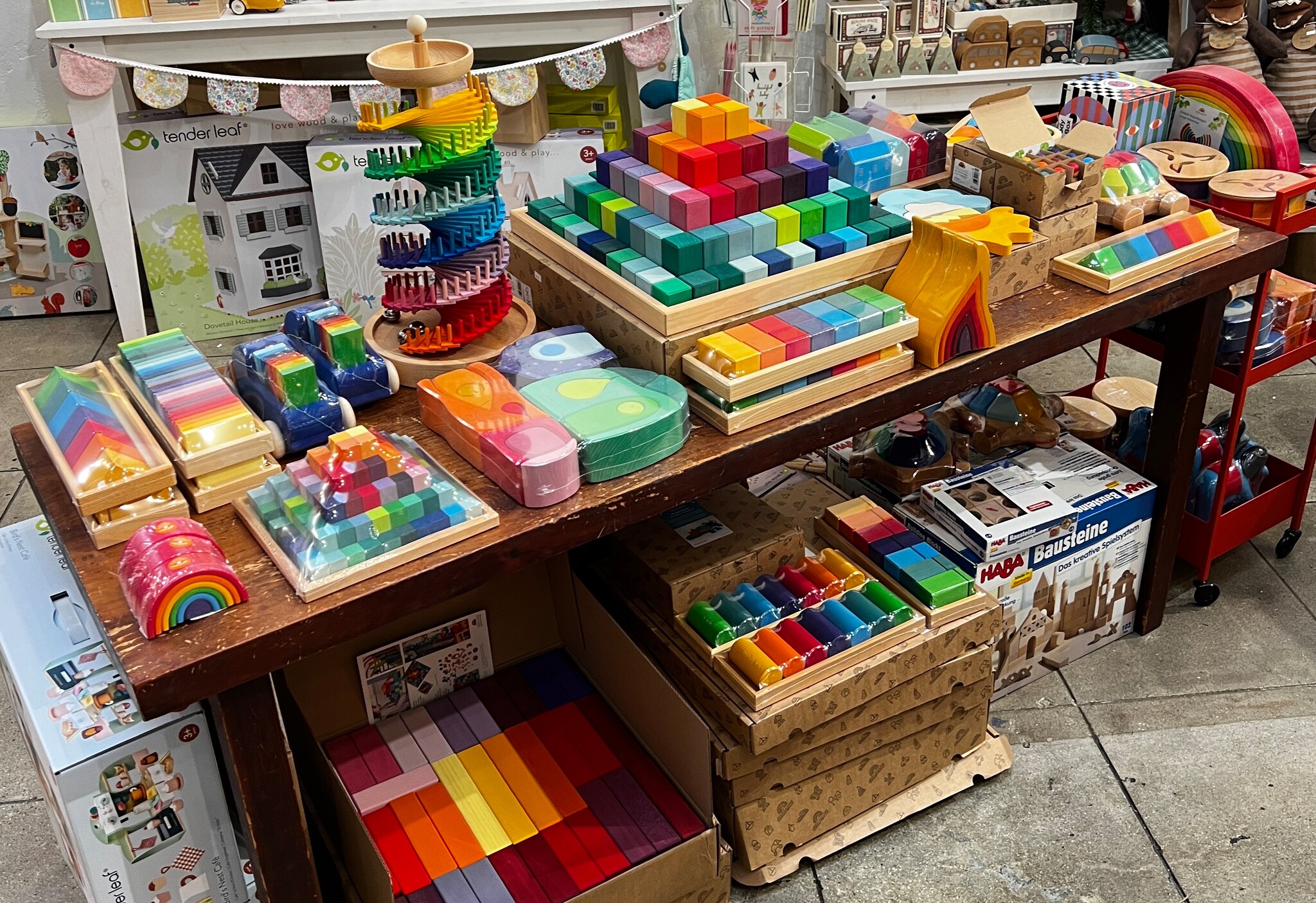 A table is packed on top and below with a rainbow of toys, including blocks, chalks, and wooden doll houses.