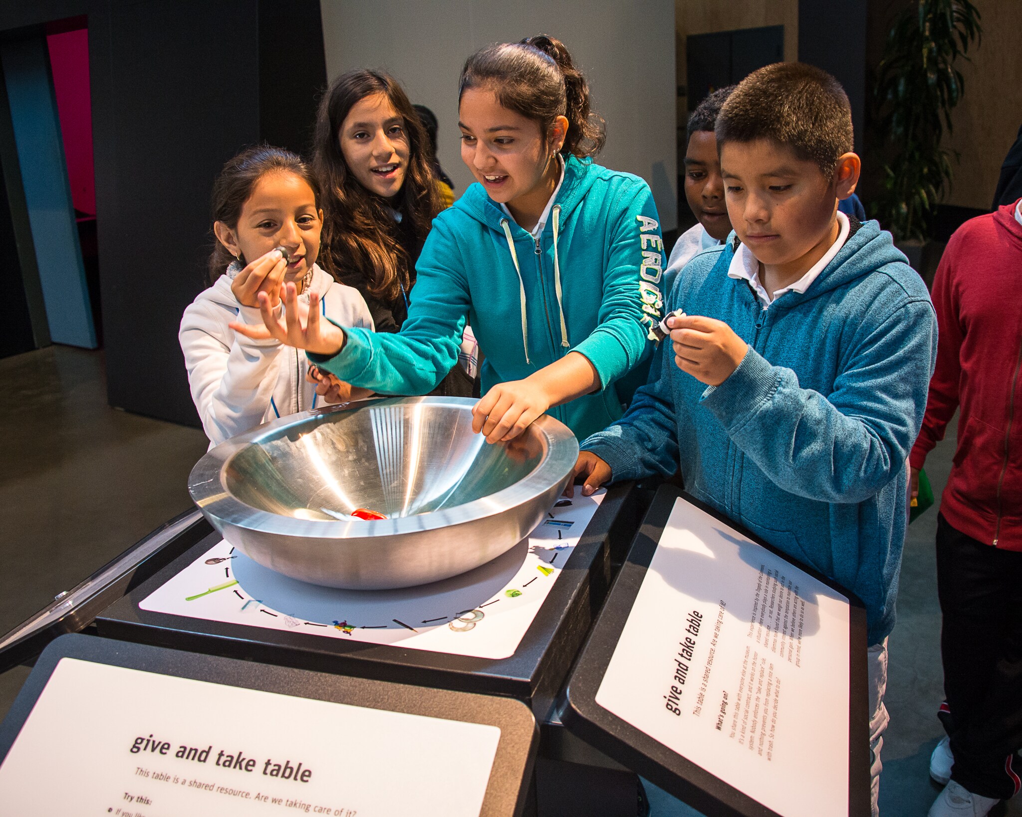 A group of middle school students of various races circle around a steel bowl on a podium with signage about sharing resources.