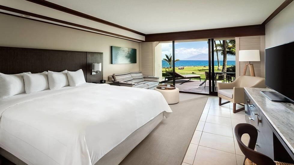 The large bed in the Oceanfront Extended Lanai guest room faces a TV. The far side of the room has seating and glass doors to the lanai.