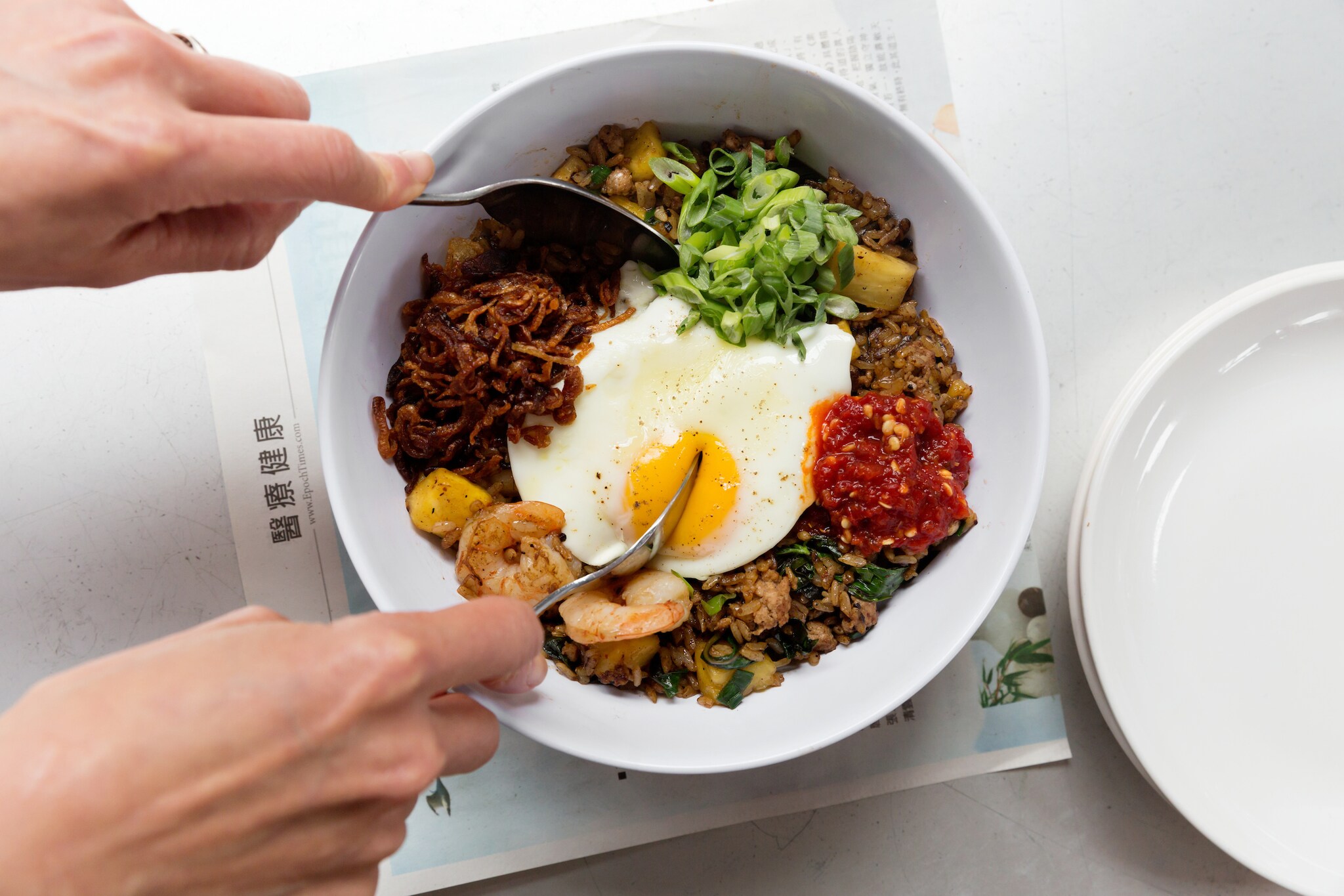 Overhead view of a pair of pale hands using a pair of spoons to dig into the fried egg atop a bowl of fried rice.