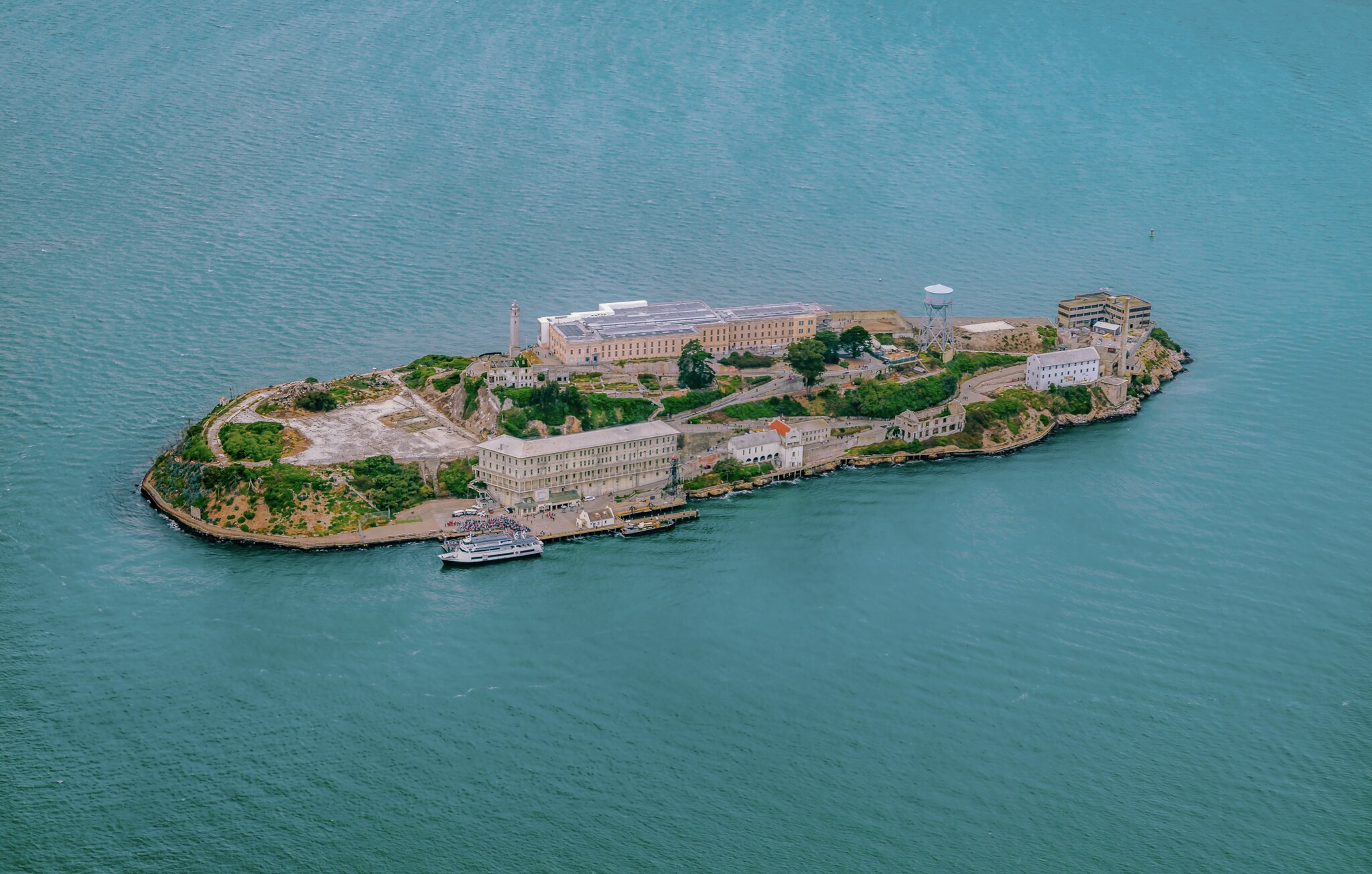 Aerial view of Alcatraz Island, a flat oval island with patches of greenery around the large rectangular buildings of the old prison complex.