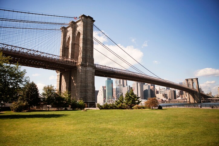 View of Brooklyn Bridge and Manhattan beyond during a sunny day at the park, with wide open green spaces and clumps of trees.