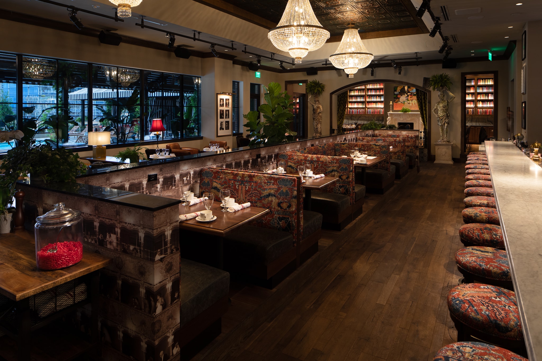 A large restaurant hosts booths with patterned upholstery, stools lining a long bard, and tall bookcases around a fireplace in the distance.